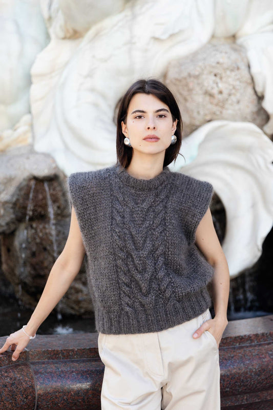 A woman wearing a brown mohair sweater vest and cream trousers, standing in front of a large white stone fountain.