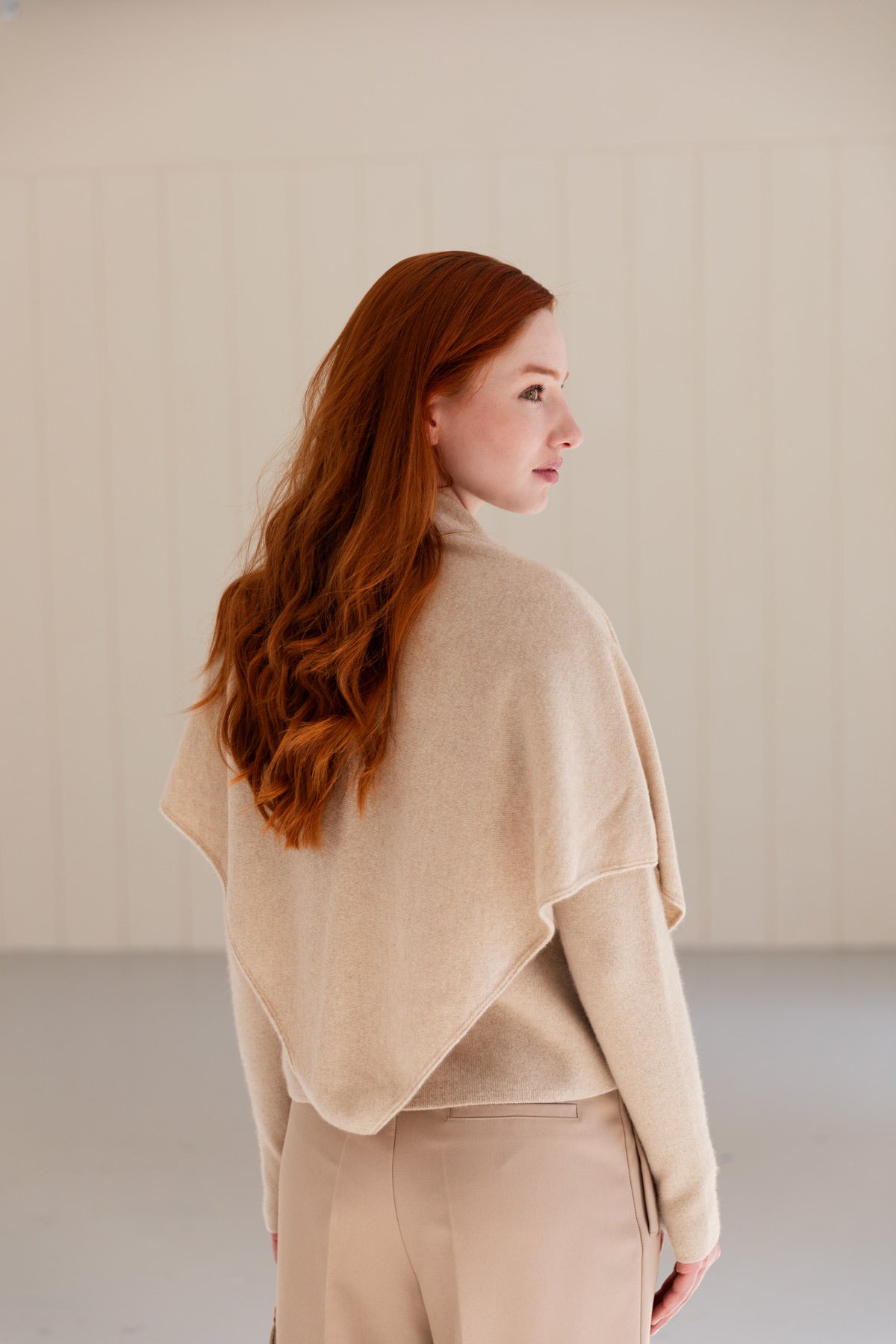 A woman is shown from the back wearing a beige cashmere triangle scarf and matching sweater and trousers.