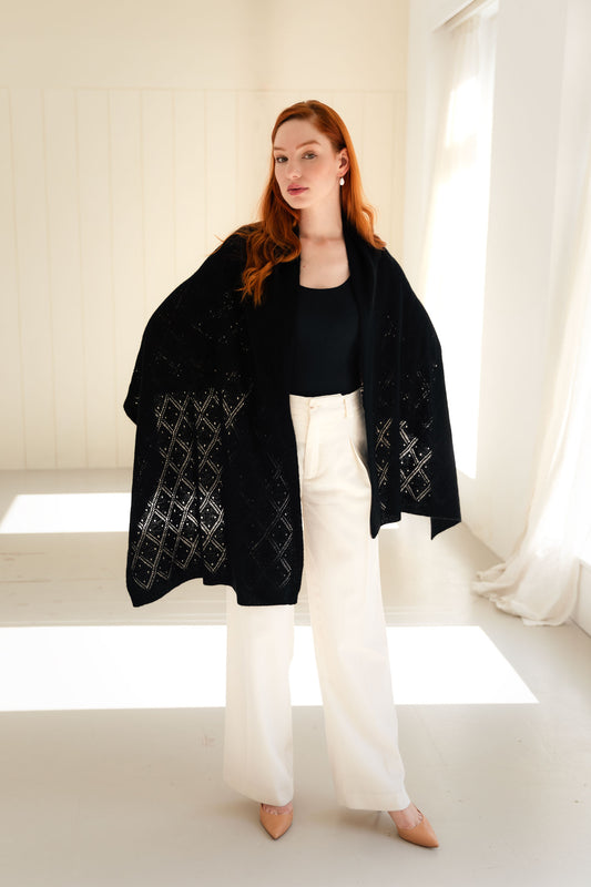 Woman wearing a black lace cashmere shawl over a black top and white pants in a bright room.