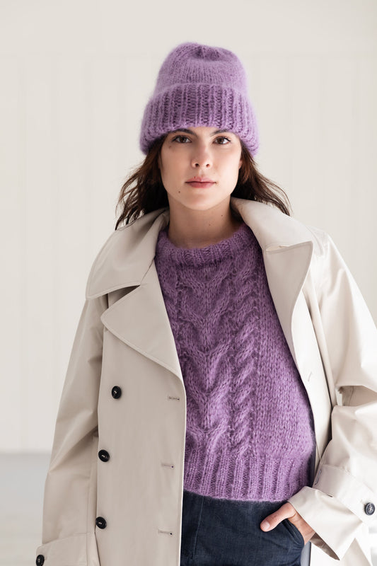 A woman wearing a lavender cable-knit mohair sweater, a matching mohair beanie, and a light beige trench coat, photographed against a clean off-white studio background.
