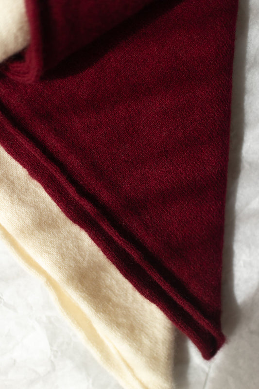 A close-up of two folded cashmere triangle scarves, showing a deep burgundy red outer layer and a cream inner layer on a light textured background.