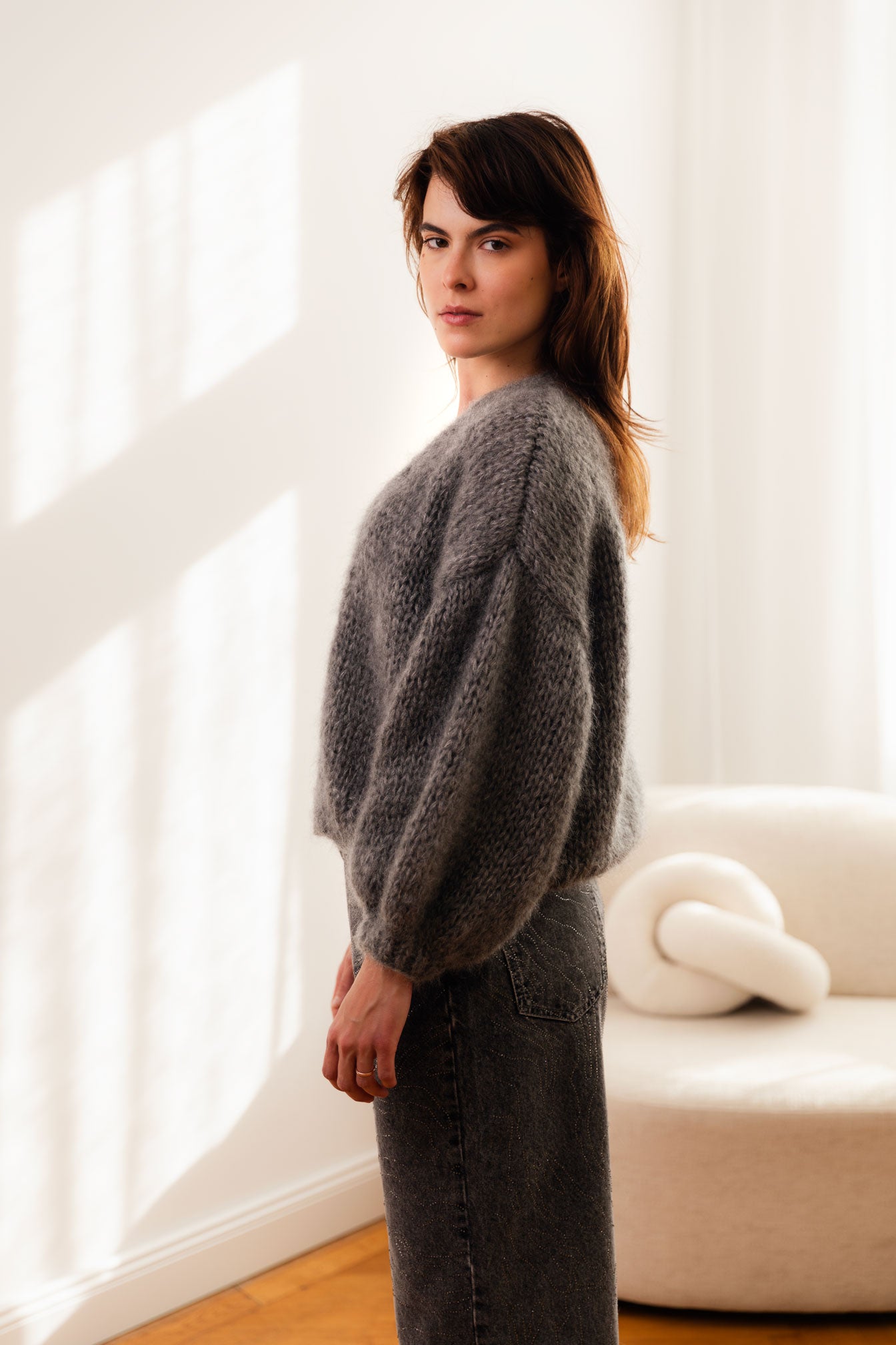 Woman wearing a grey mohair cardigan and dark jeans standing in a room with white curtains.