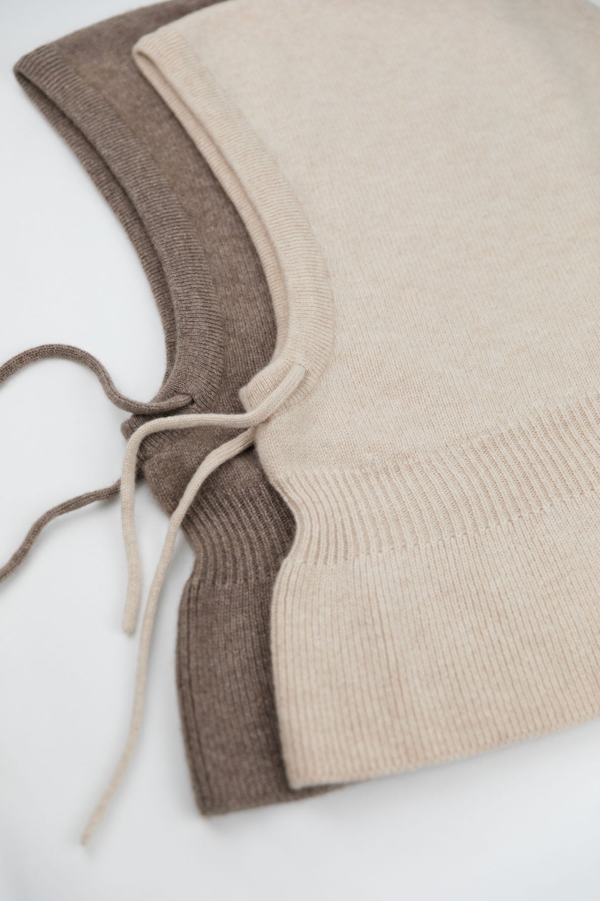 Detailed view of two 100% Mongolian cashmere balaclavas in beige and brown, showing the adjustable drawstring and wide shoulder drape.