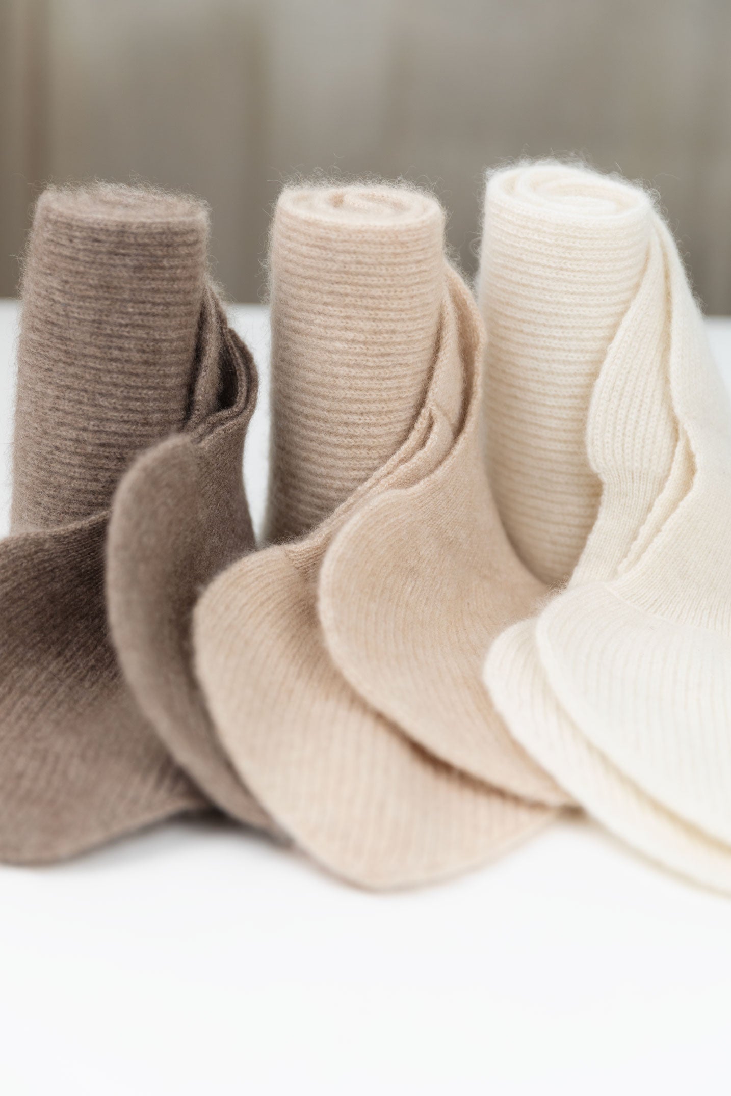 Three rolled cashmere accessories in Taupe, Oatmeal, and Natural White, showcasing the soft texture of undyed Mongolian cashmere fibers.