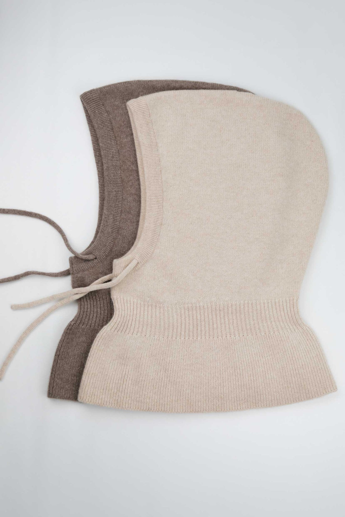Side view of two 100% Mongolian cashmere balaclavas in beige and brown, showing the adjustable drawstring and wide shoulder drape.