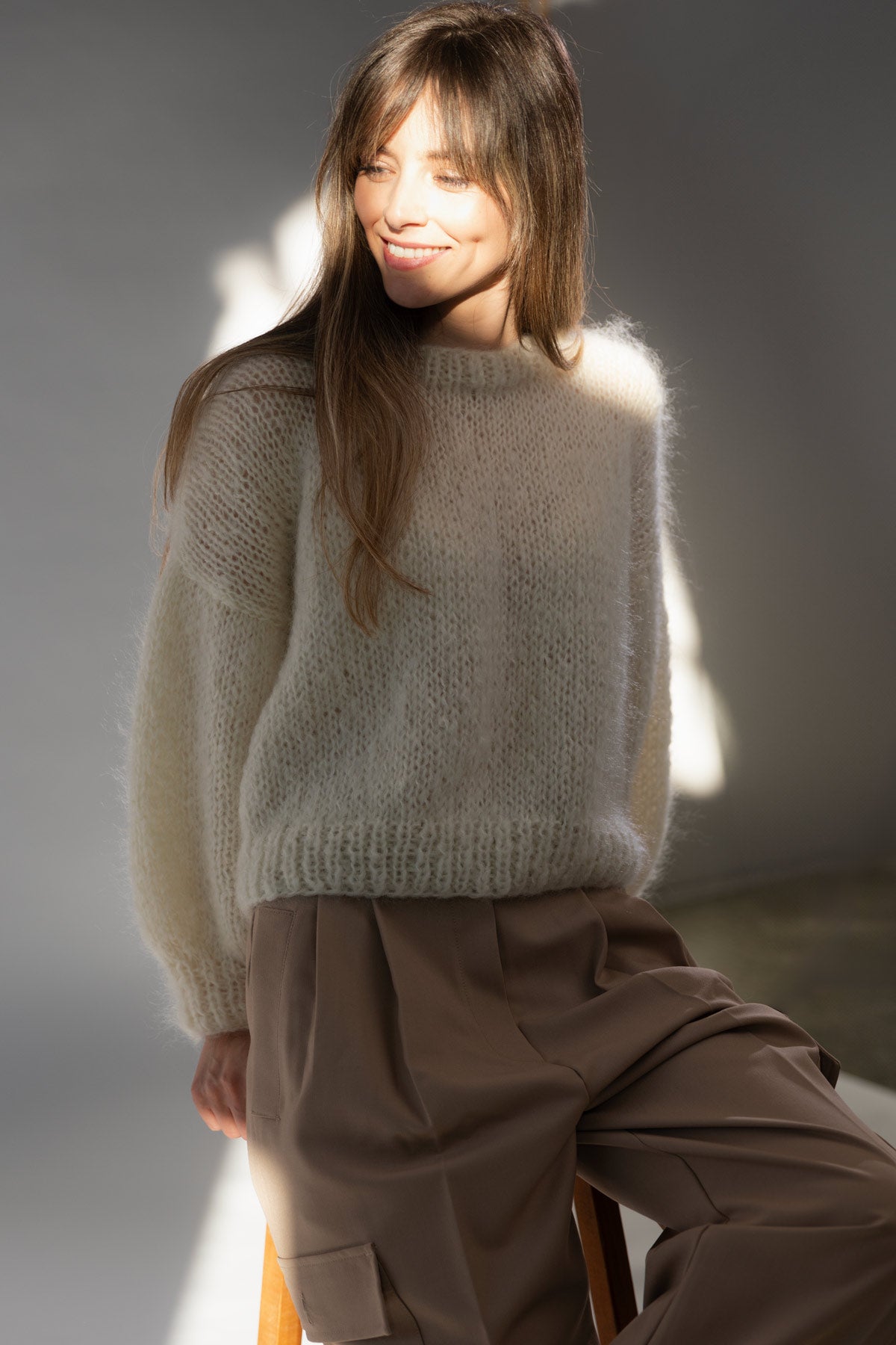 A woman wearing a fuzzy off-white mohair sweater and brown wide-leg pants, seated in a softly lit studio setting.