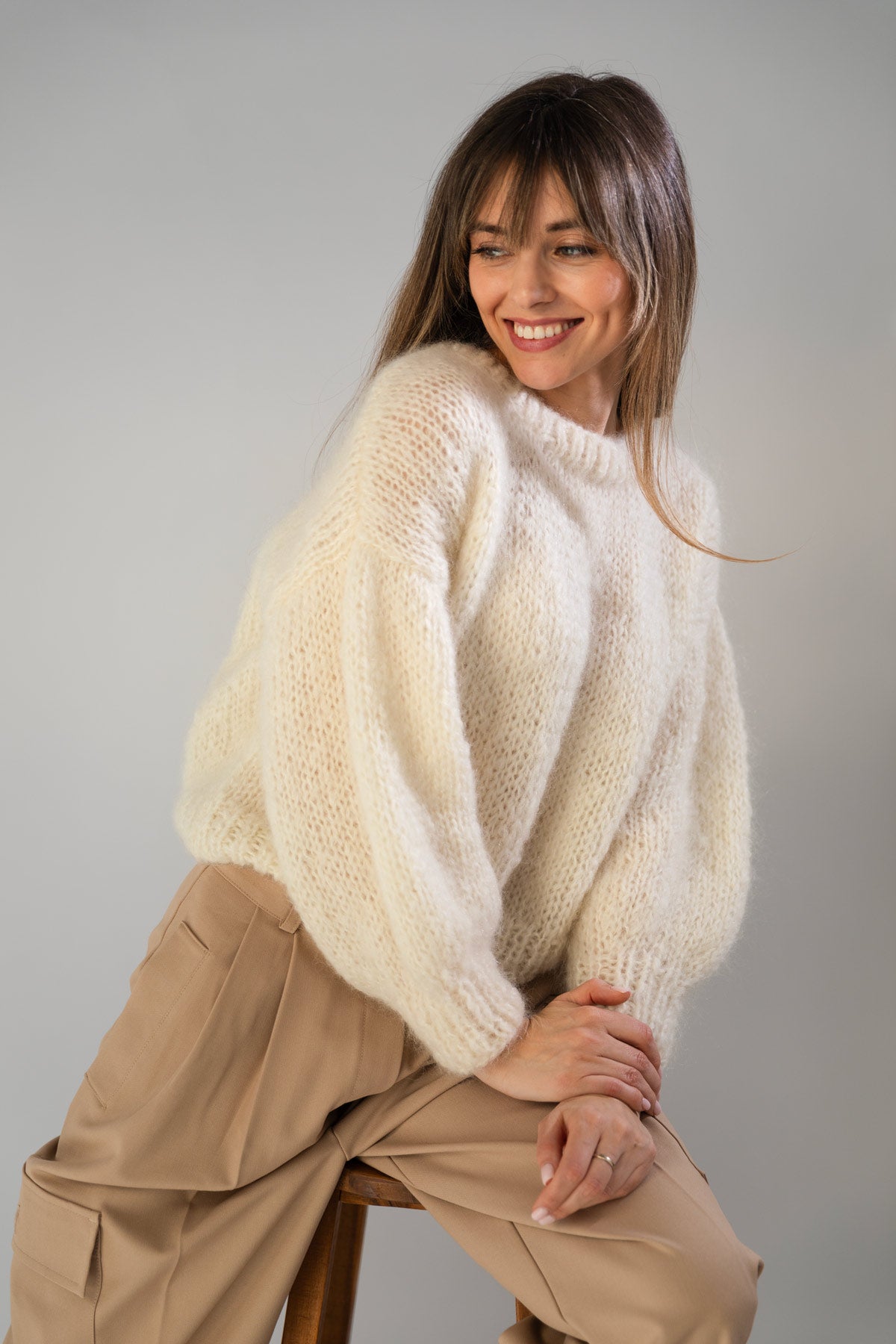 A woman wearing a chunky, oversized cream-colored mohair sweater and beige trousers, sitting on a wooden stool