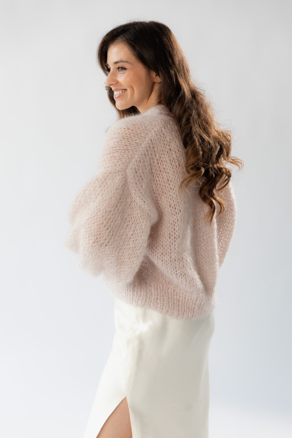 A woman with long hair is shown from the back and side, wearing an elegant beige mohair cardigan and a white silk dress against a plain light grey studio background.