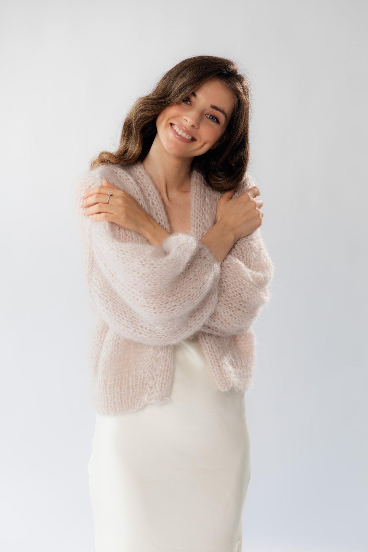 A woman wearing an airy mohair cardigan and a white silk dress, posing with her arms crossed in front of her chest against a light  studio background.