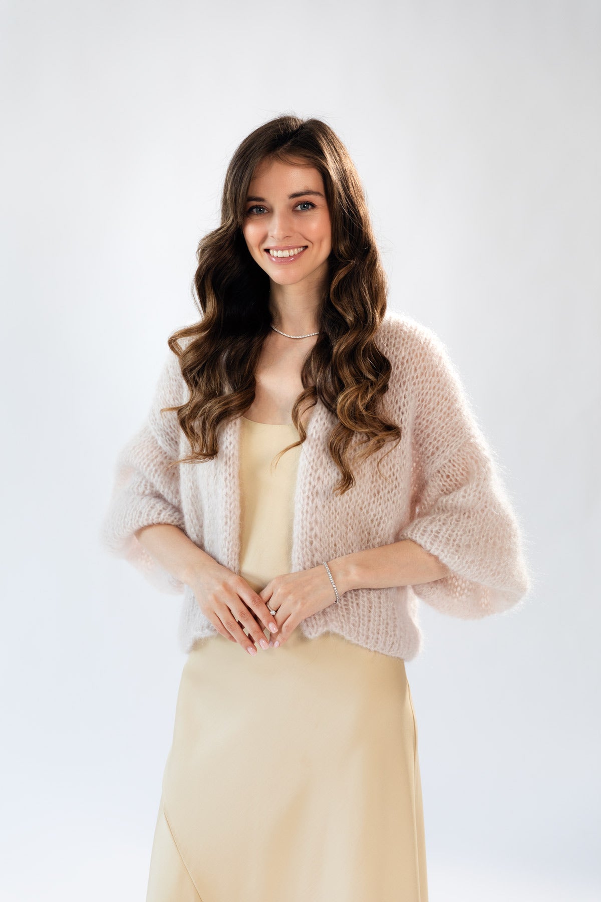 A woman with long wavy brown hair wearing a light beige mohair cardigan over a beige silk dress, set against a clean white studio background.