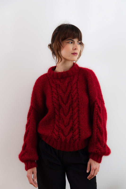 A woman wearing a deep red cable-knit mohair sweater with balloon sleeves and black pants, standing against a plain white studio background.