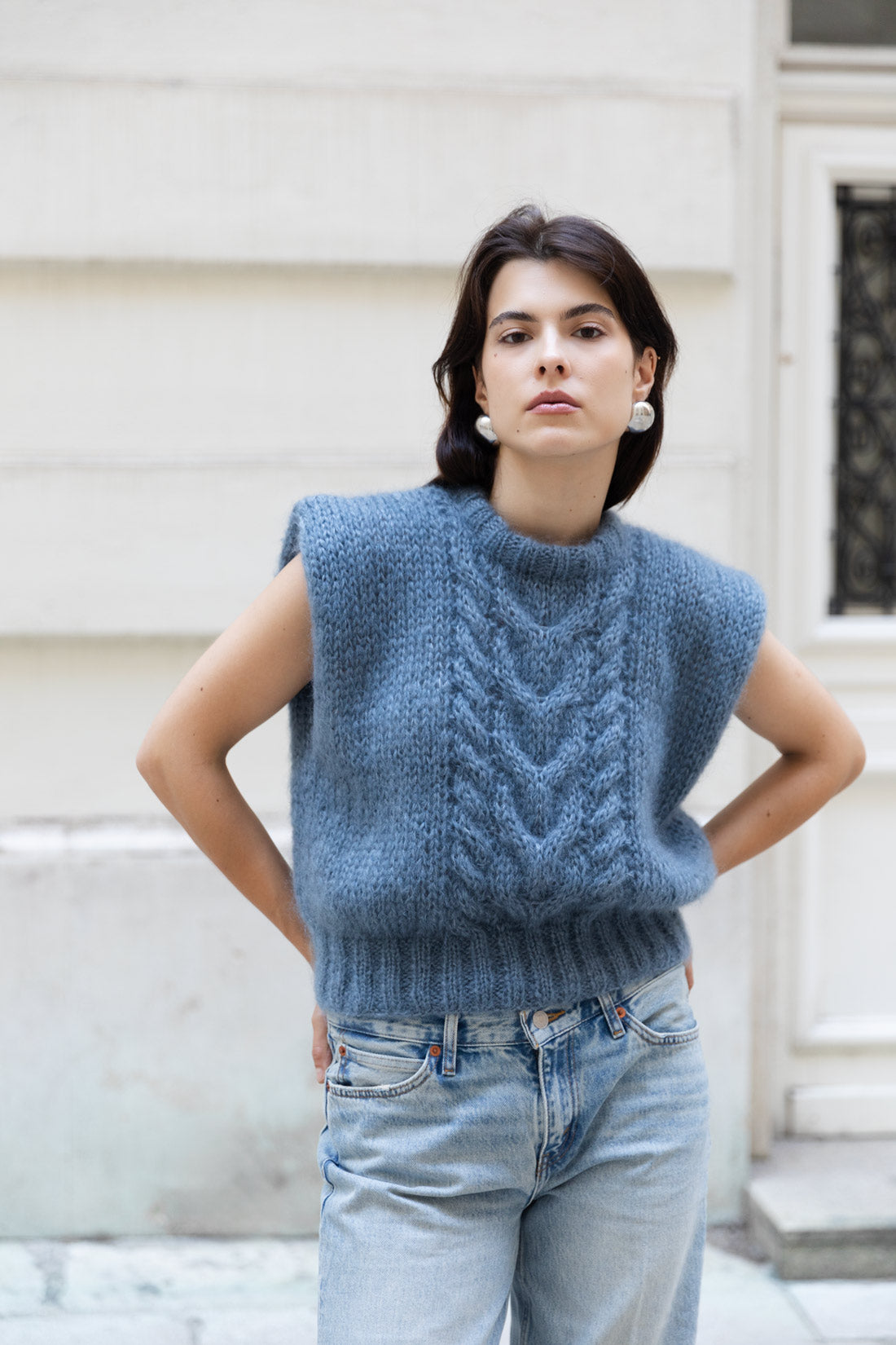 A woman wearing a sleeveless mohair cable-knit sweater vest in a dusty slate blue, paired with light-wash blue jeans, standing outdoors against a light-coloured marble wall.