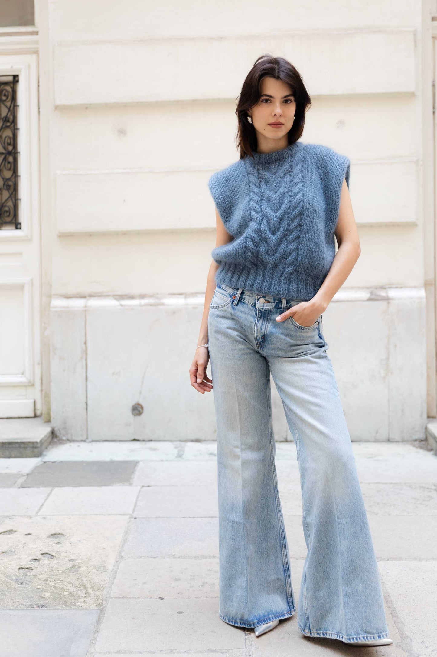Woman wearing a blue sleeveless mohair vest and light blue jeans standing against a white wall.