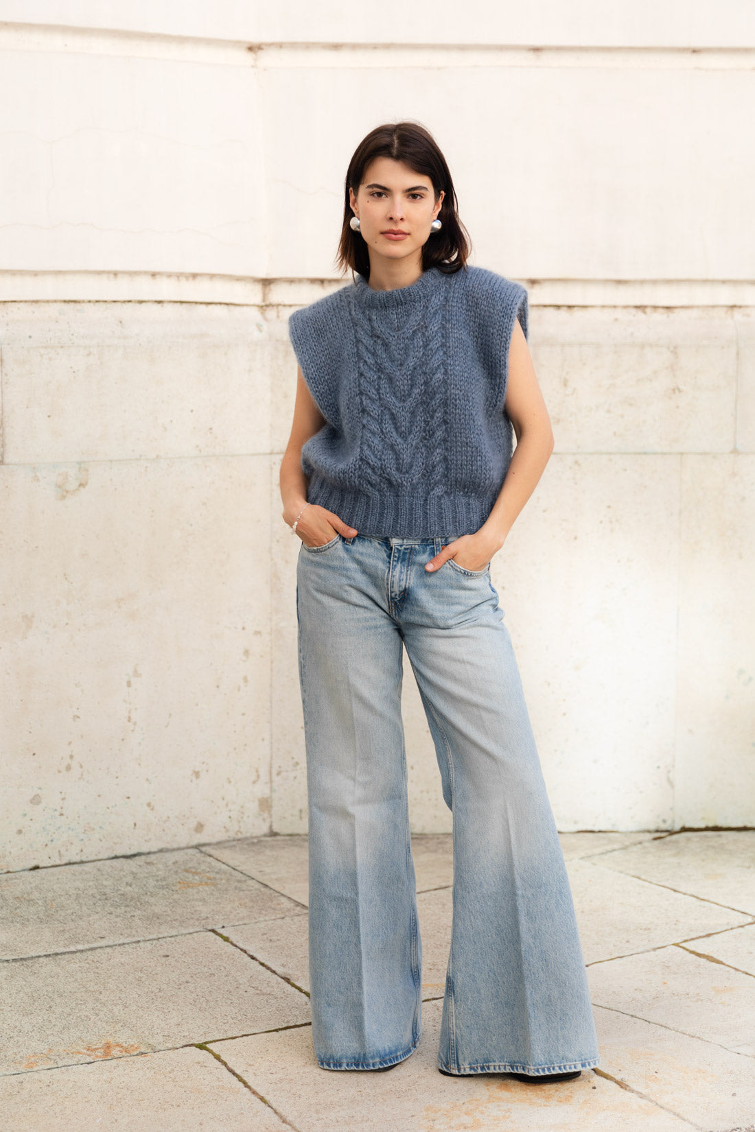 A woman wearing a sleeveless, cable-knit mohair sweater vest in blue and light-wash wide-leg denim jeans, standing against a beige stone wall.