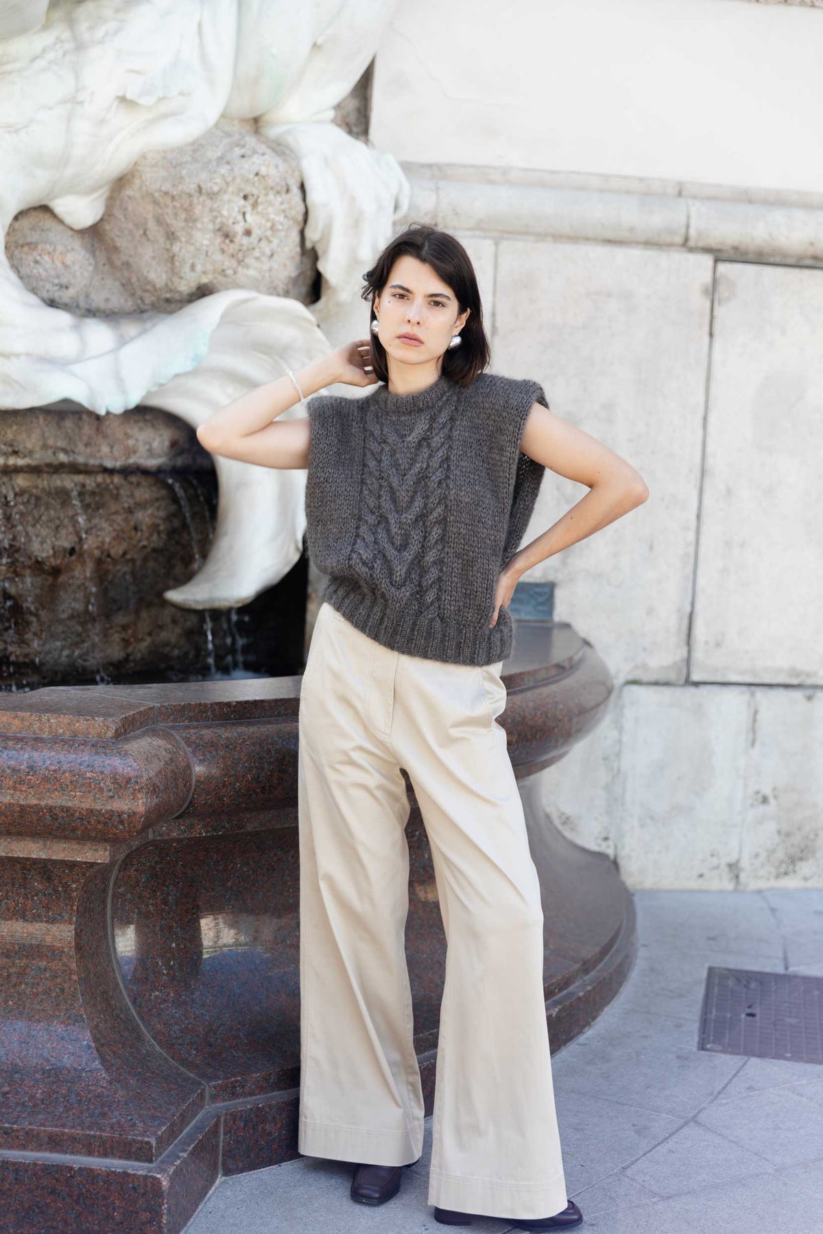Woman wearing an ash brown mohair sweater vest and beige wide-leg pants standing in front of a marble fountain.