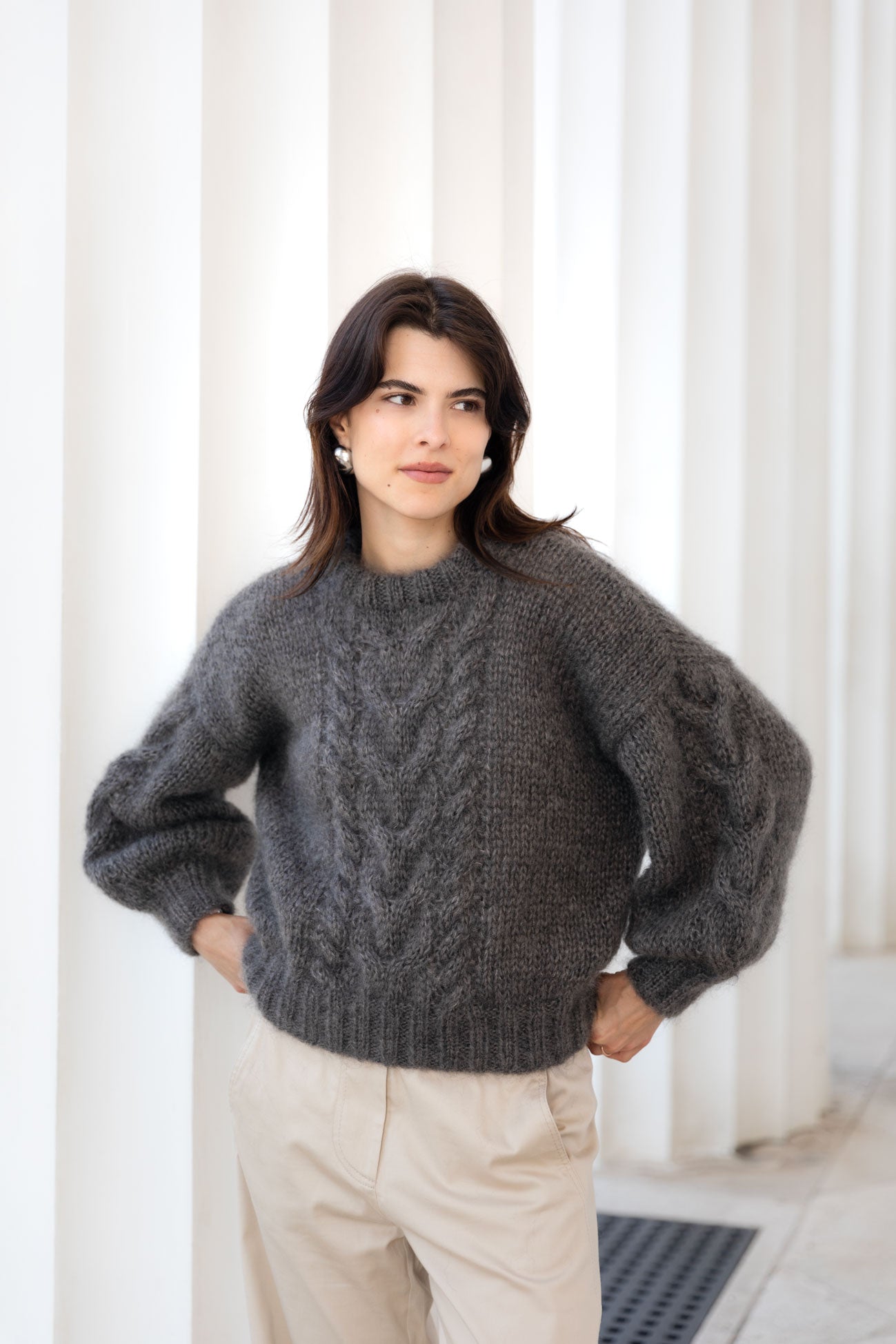 Woman wearing a brown mohair sweater, paired with beige trousers, sitting against a white background