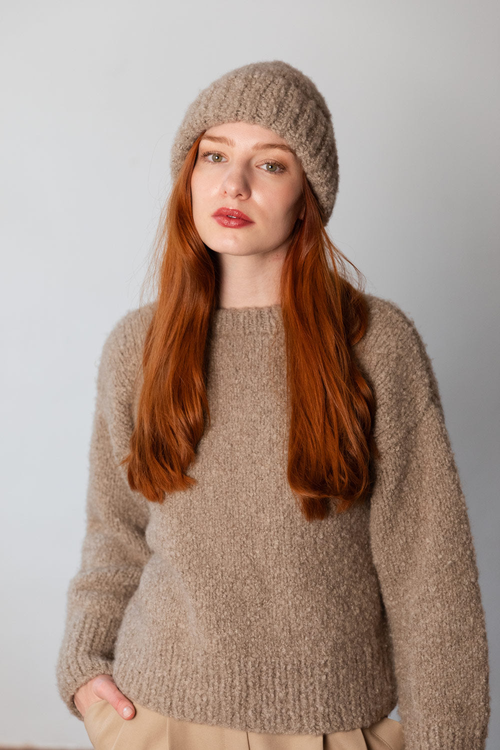 A woman with long red hair wearing a chunky beige cashmere sweater and matching beanie, photographed against a plain light grey background.