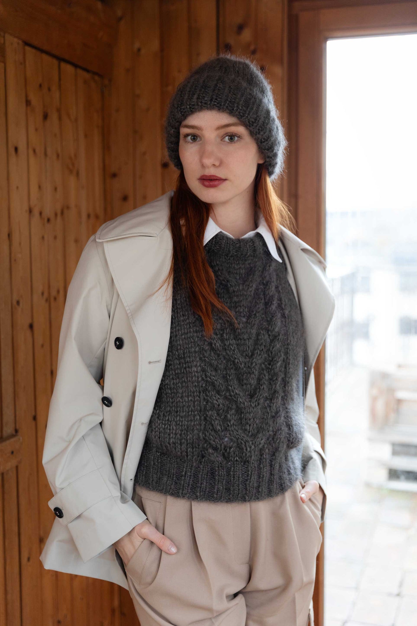 A woman wearing a dark gray cable-knit sweater vest, a matching gray mohair beanie, a light beige trench coat, and beige trousers, standing in a rustic wooden interior.