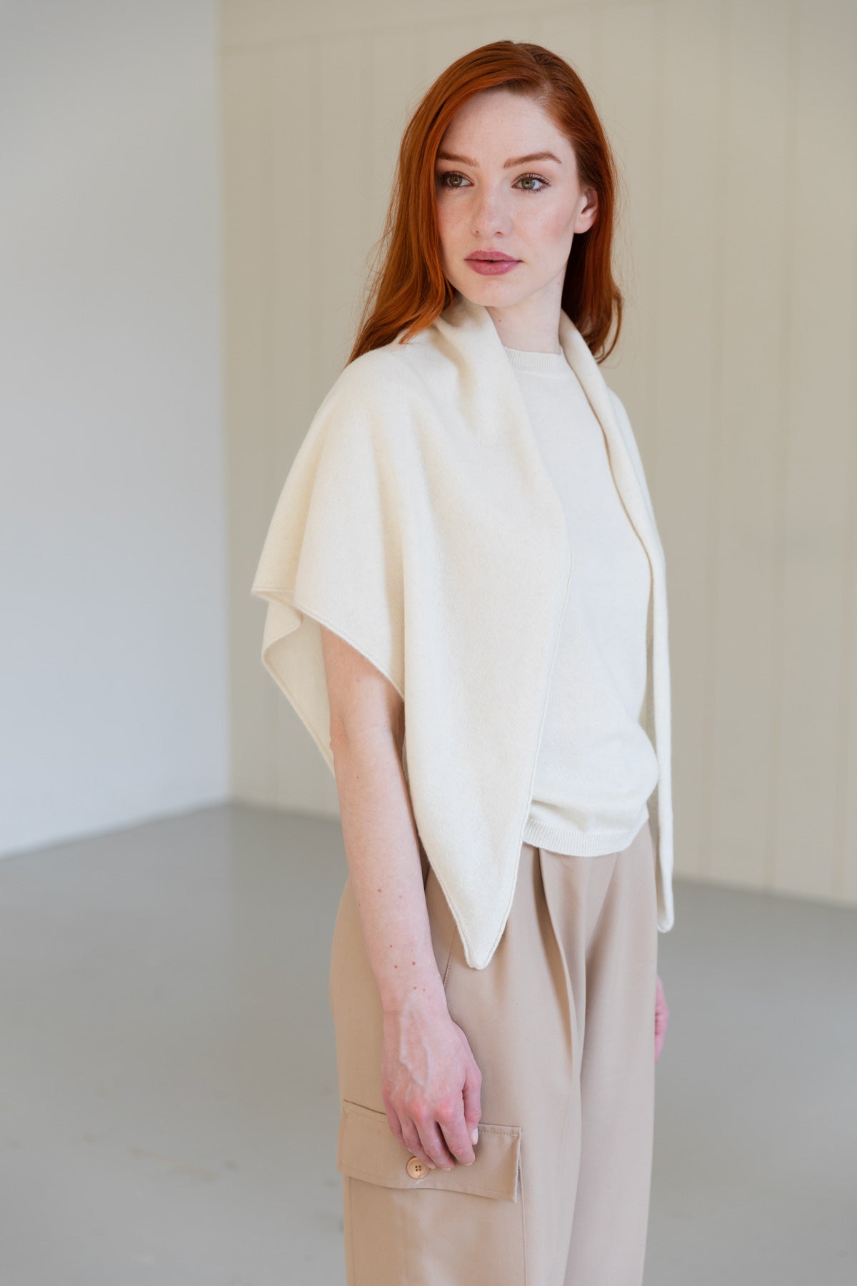 A woman with red hair wearing a cream cashmere triangle scarf, paired with a white cashmere t-shirt, and beige trousers.