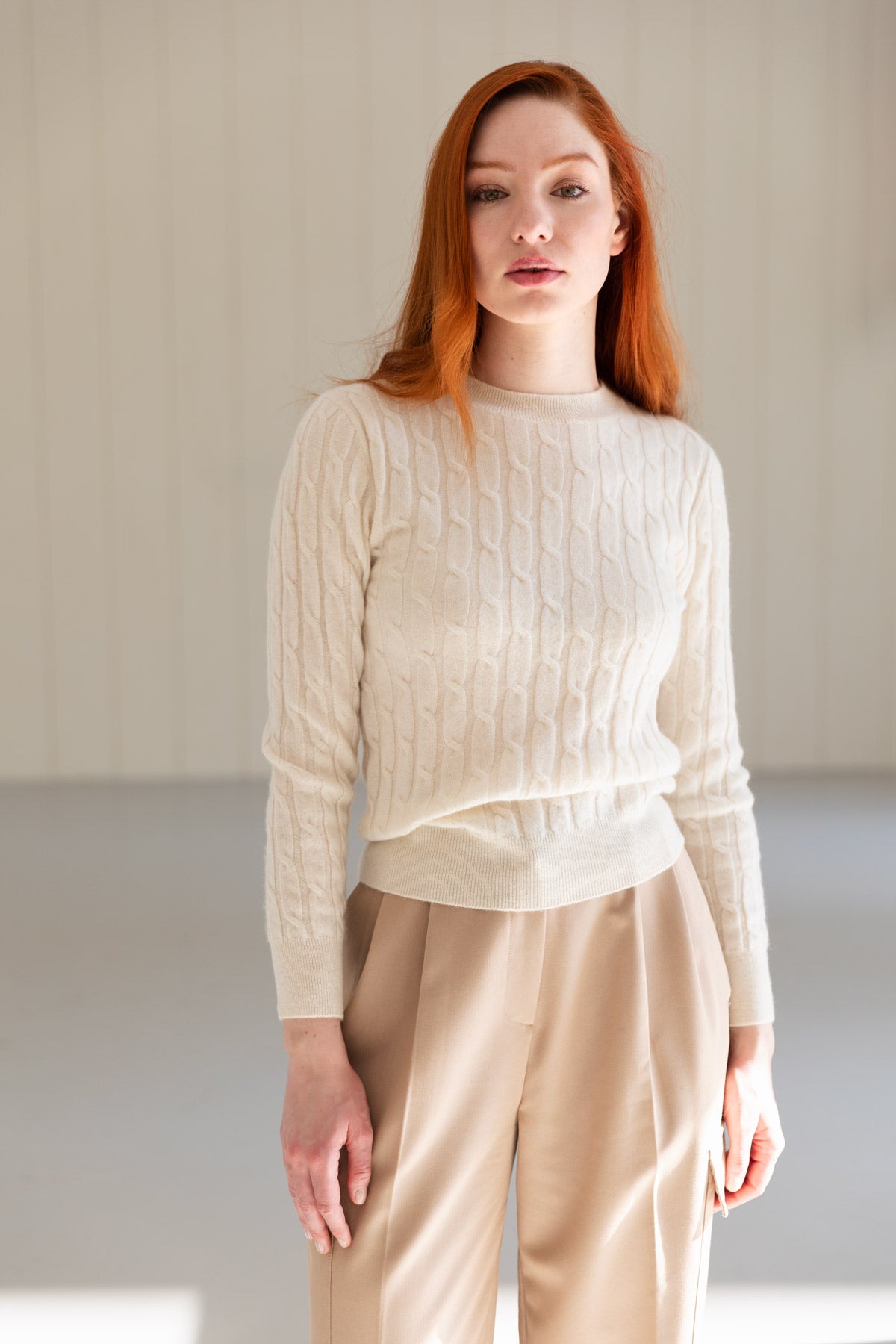 A woman with red hair wearing a cream cable-knit cashmere sweater and beige trousers, standing in a minimalist room.