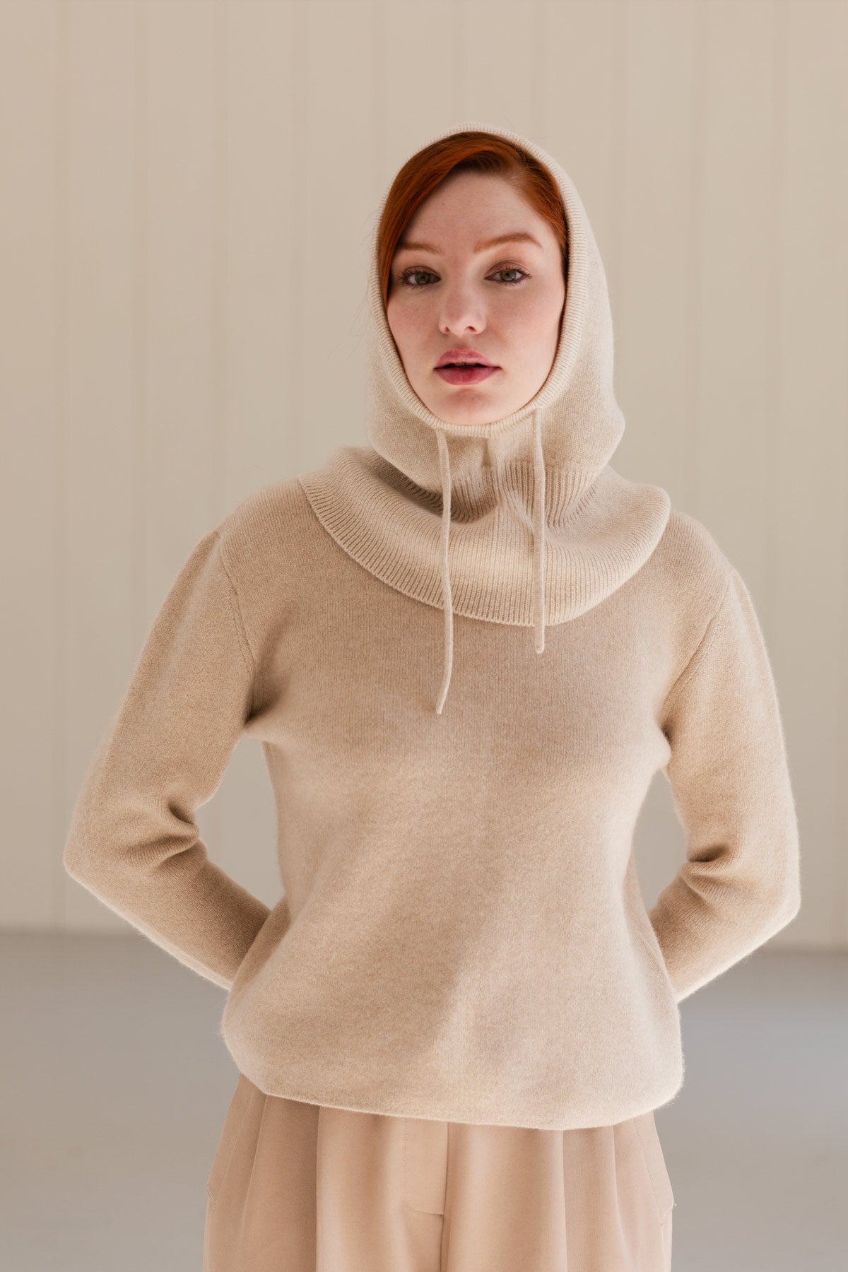 A woman wearing a beige cashmere balaclava over a matching cashmere sweater and coordinating beige pants, standing against a light beige panelled wall.