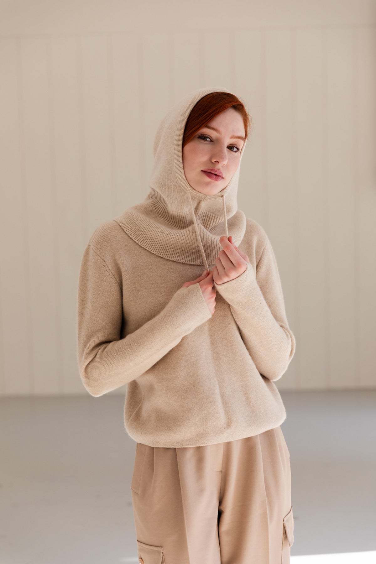 A woman wearing a beige cashmere balaclava over a matching cashmere sweater and coordinating beige pants, standing against a light beige panelled wall.