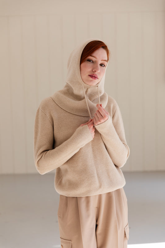A woman wearing a beige cashmere balaclava over a matching cashmere sweater and coordinating beige pants, standing against a light beige panelled wall.