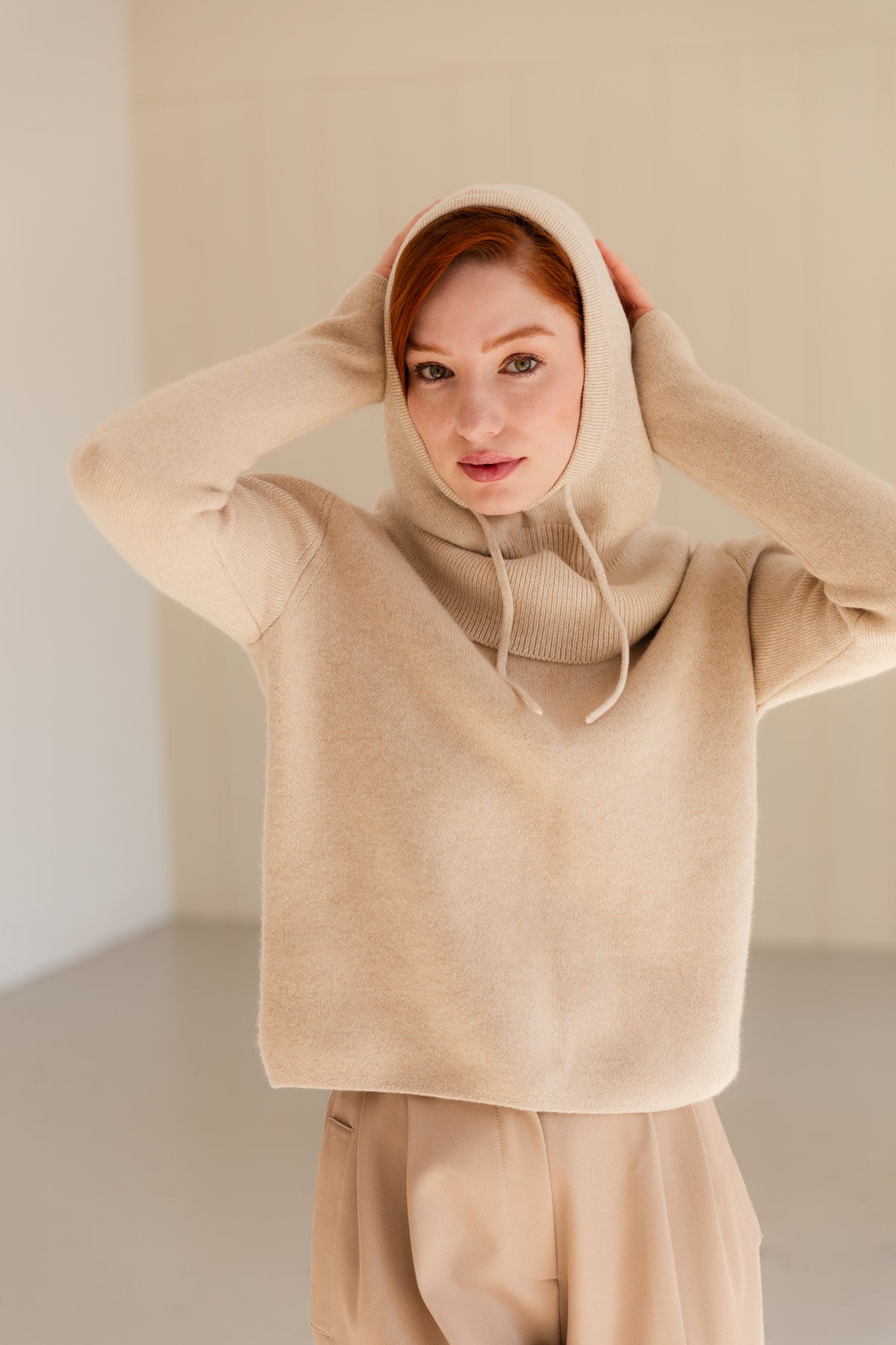 A woman wearing a beige cashmere balaclava over a matching sweater and matching beige pants, standing in a softly lit neutral indoor setting.