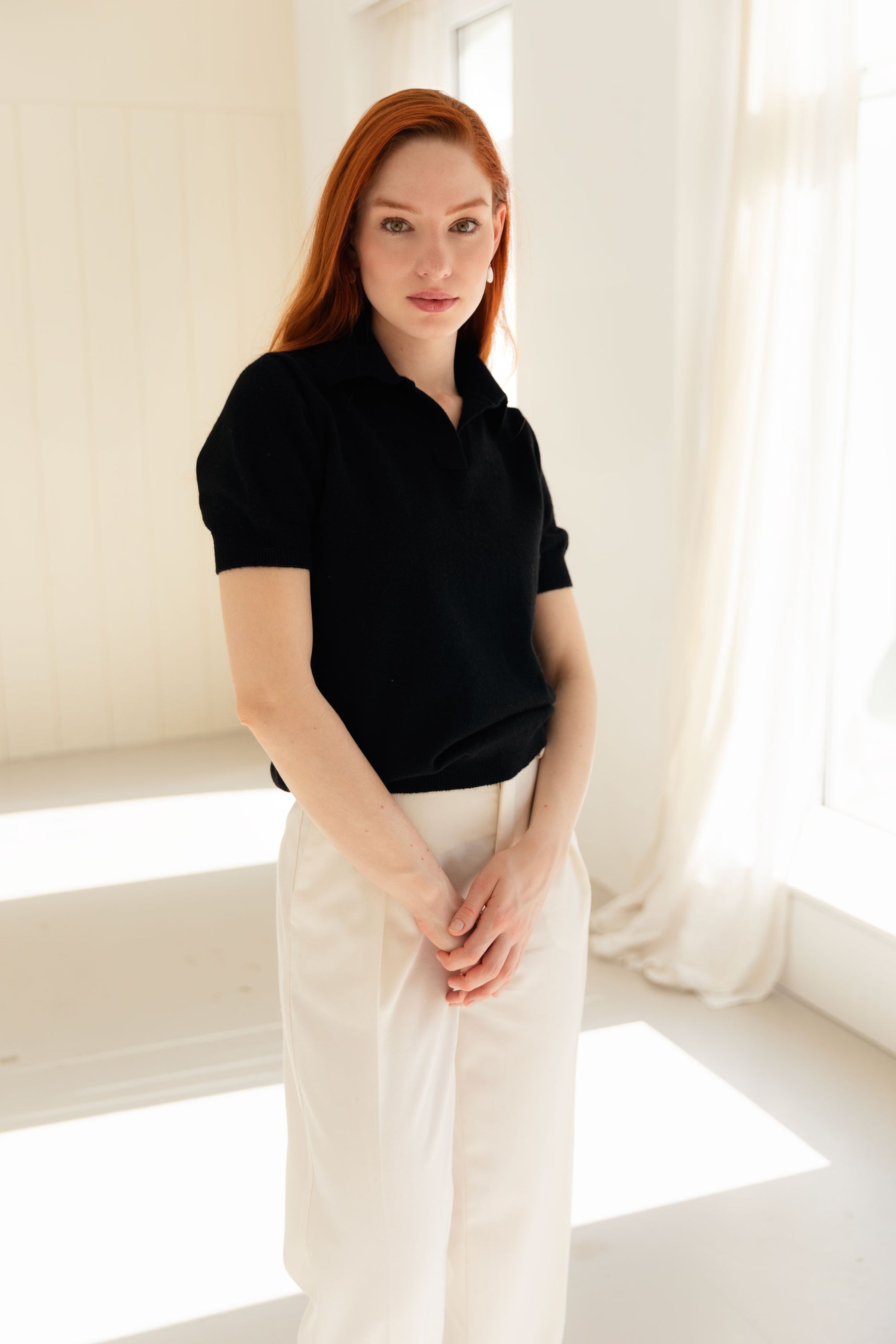 Woman wearing a black cashmere polo shirt and white pants in a bright room.