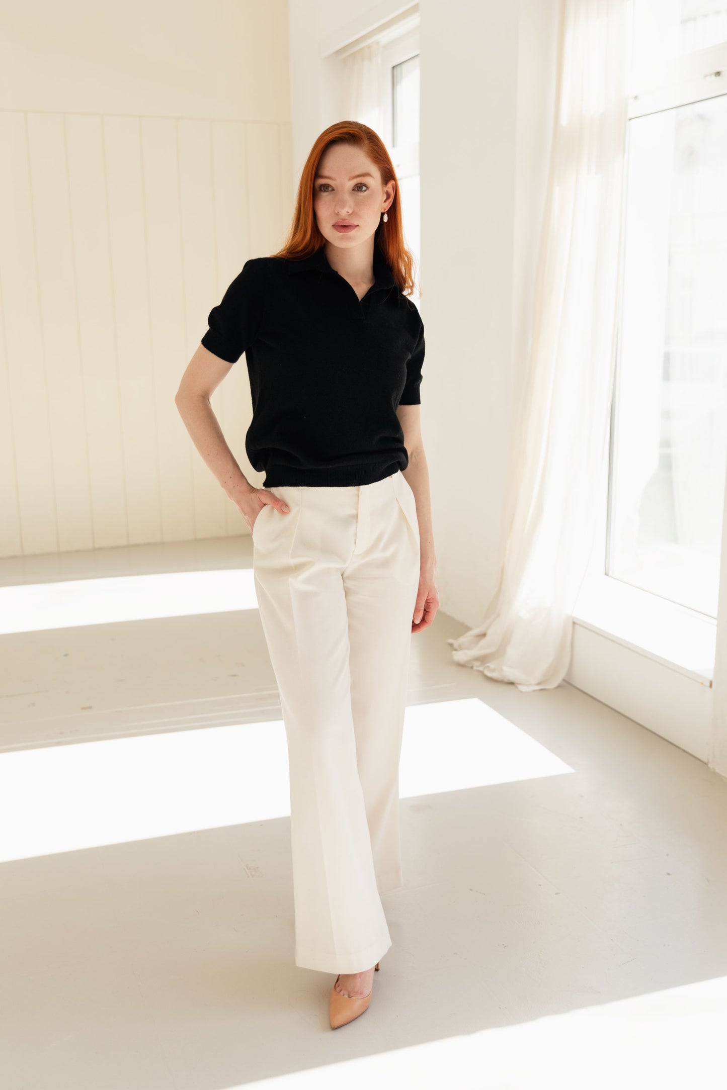 Woman wearing a black cashmere polo shirt and white pants in a bright room with large windows.
