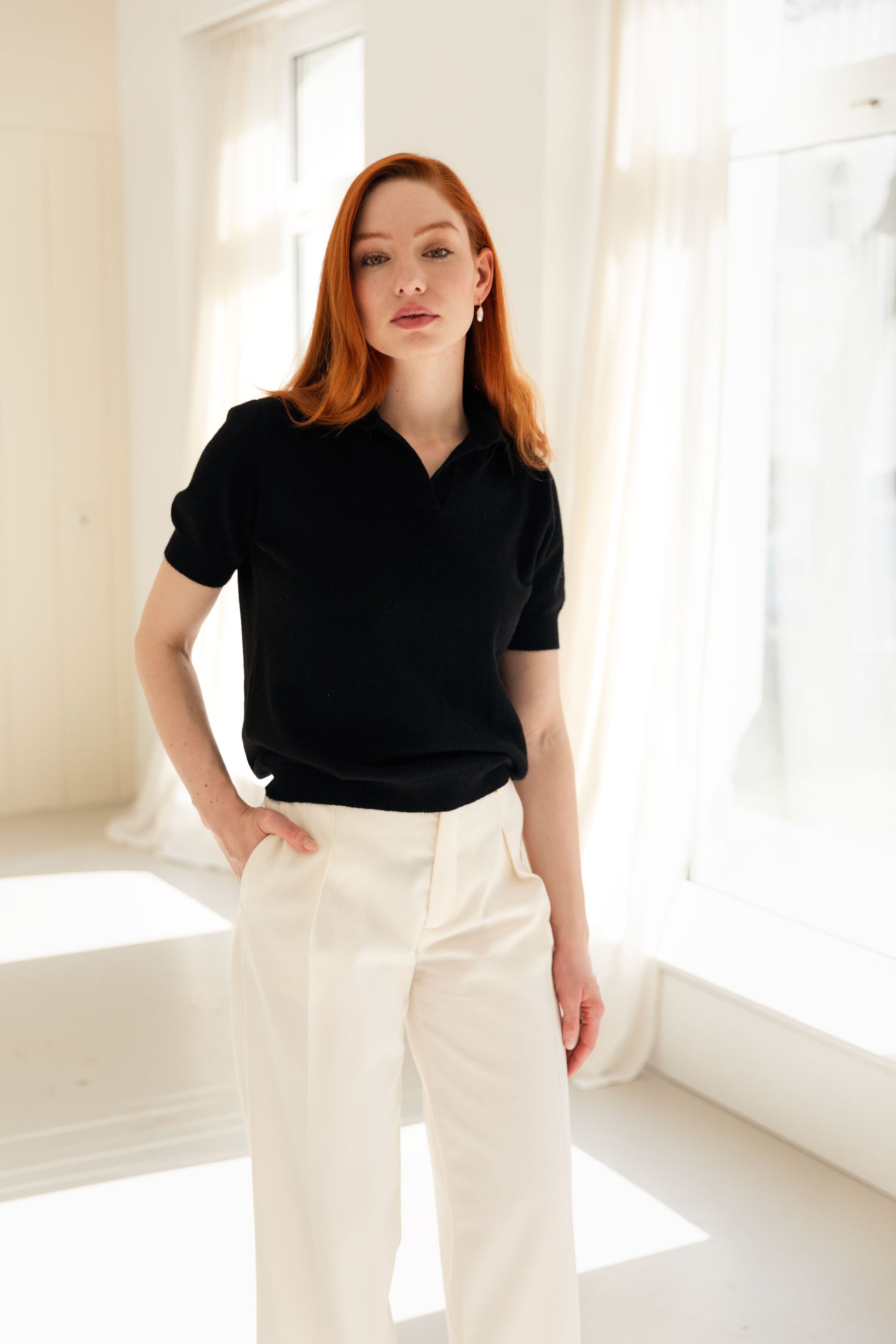 Woman wearing a black cashmere polo shirt and white pants in a bright room.