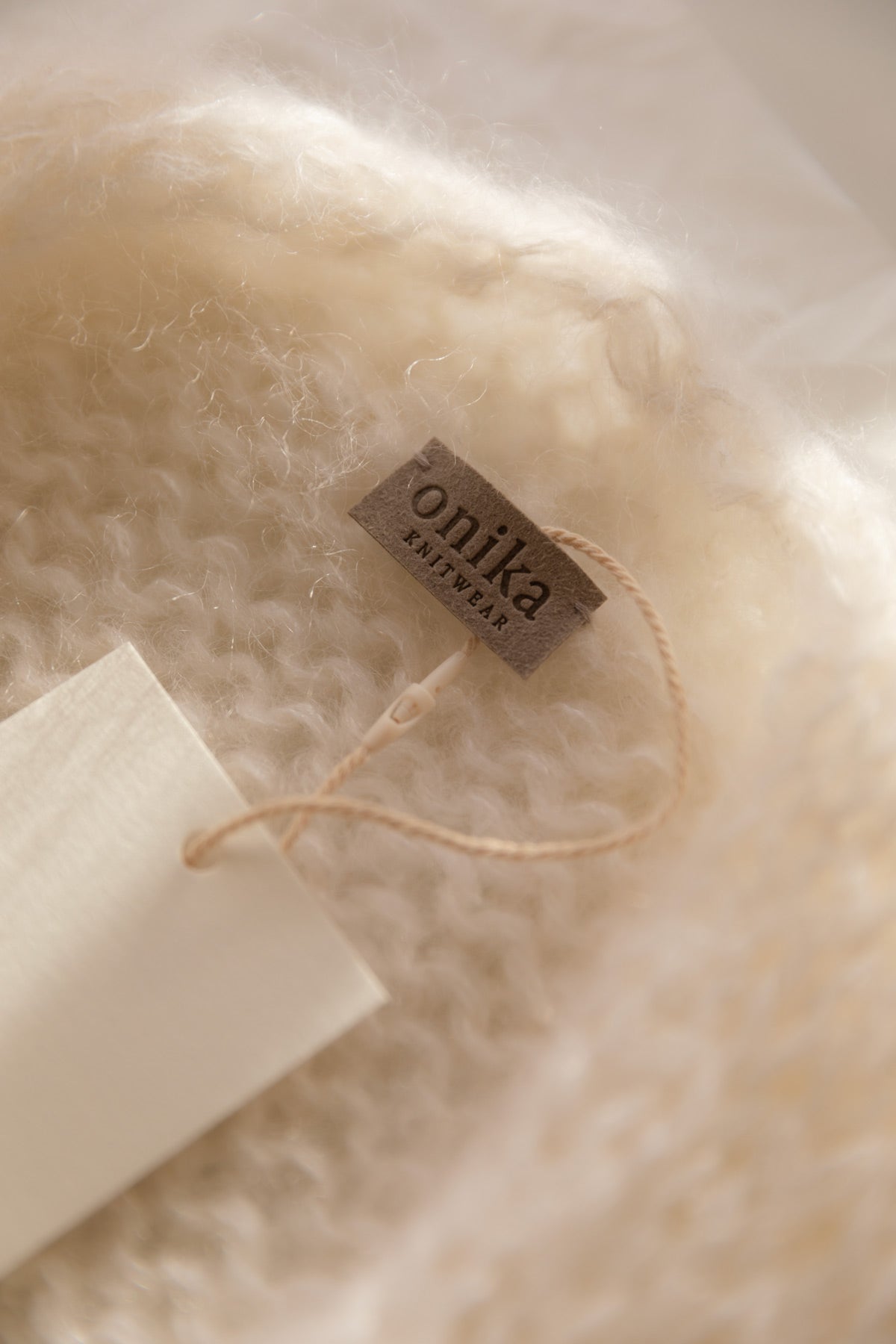 Close-up of a soft, cream-colored mohair knit garment with a small brown brand tag reading 'onika knitwear' attached by a thin string.