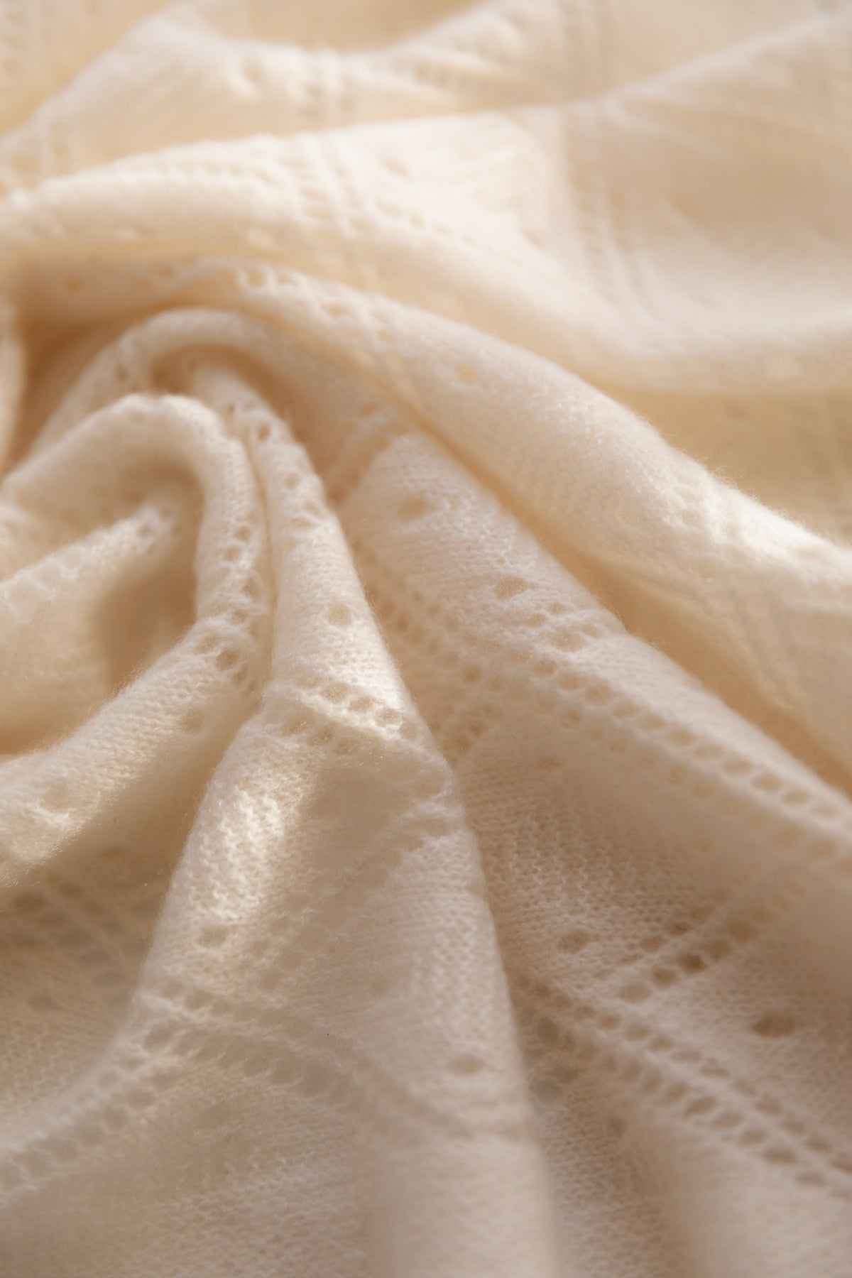 A close-up of draped cream-coloured cashmere fabric with a delicate eyelet lace pattern, showing soft folds and texture.