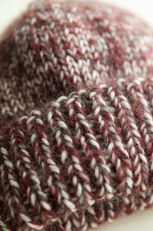 A close-up of a chunky knit beanie hat made from wool in burgundy and white, showing the detailed knit texture.