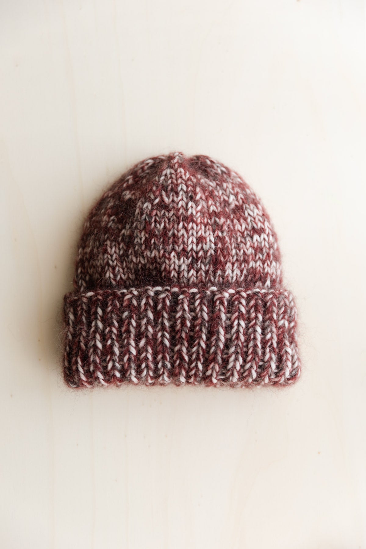 A hand-knit beanie hat in a marled mix of burgundy, brown, and cream wool, photographed on a warm off-white surface.