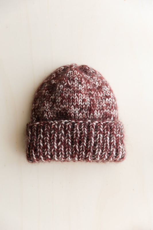 A hand-knit beanie hat in a marled mix of burgundy, brown, and cream wool, photographed on a warm off-white surface.