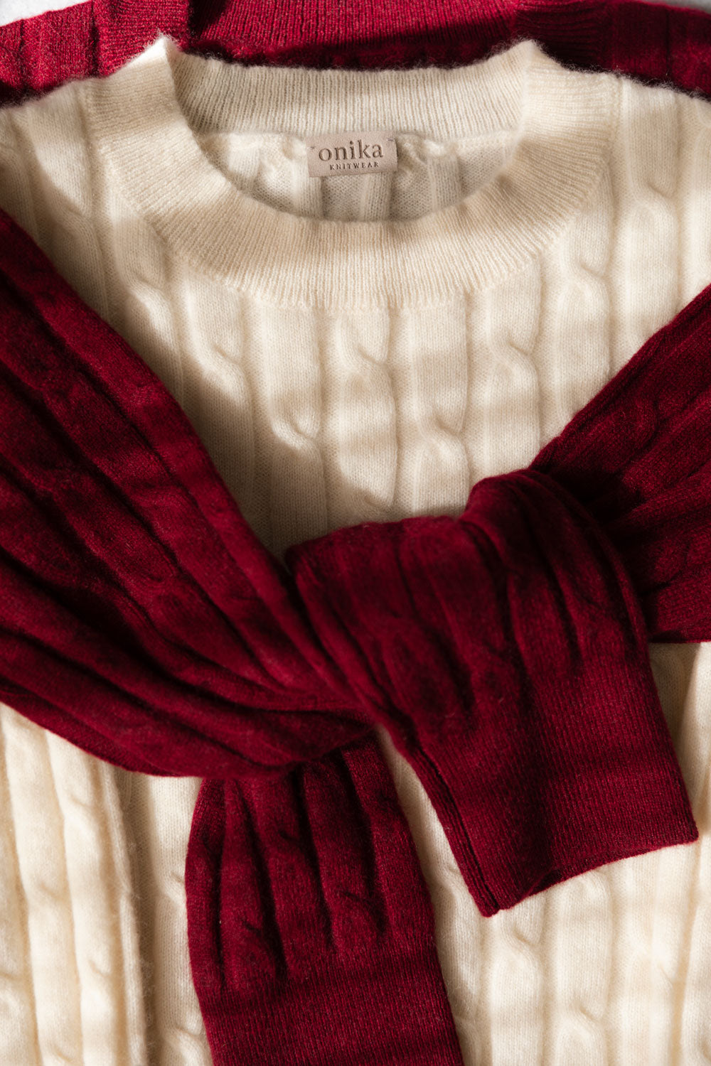 A close-up of two cable-knit cashmere sweaters, in deep burgundy and cream, showing the brand label 'onika knitwear' at the collar.