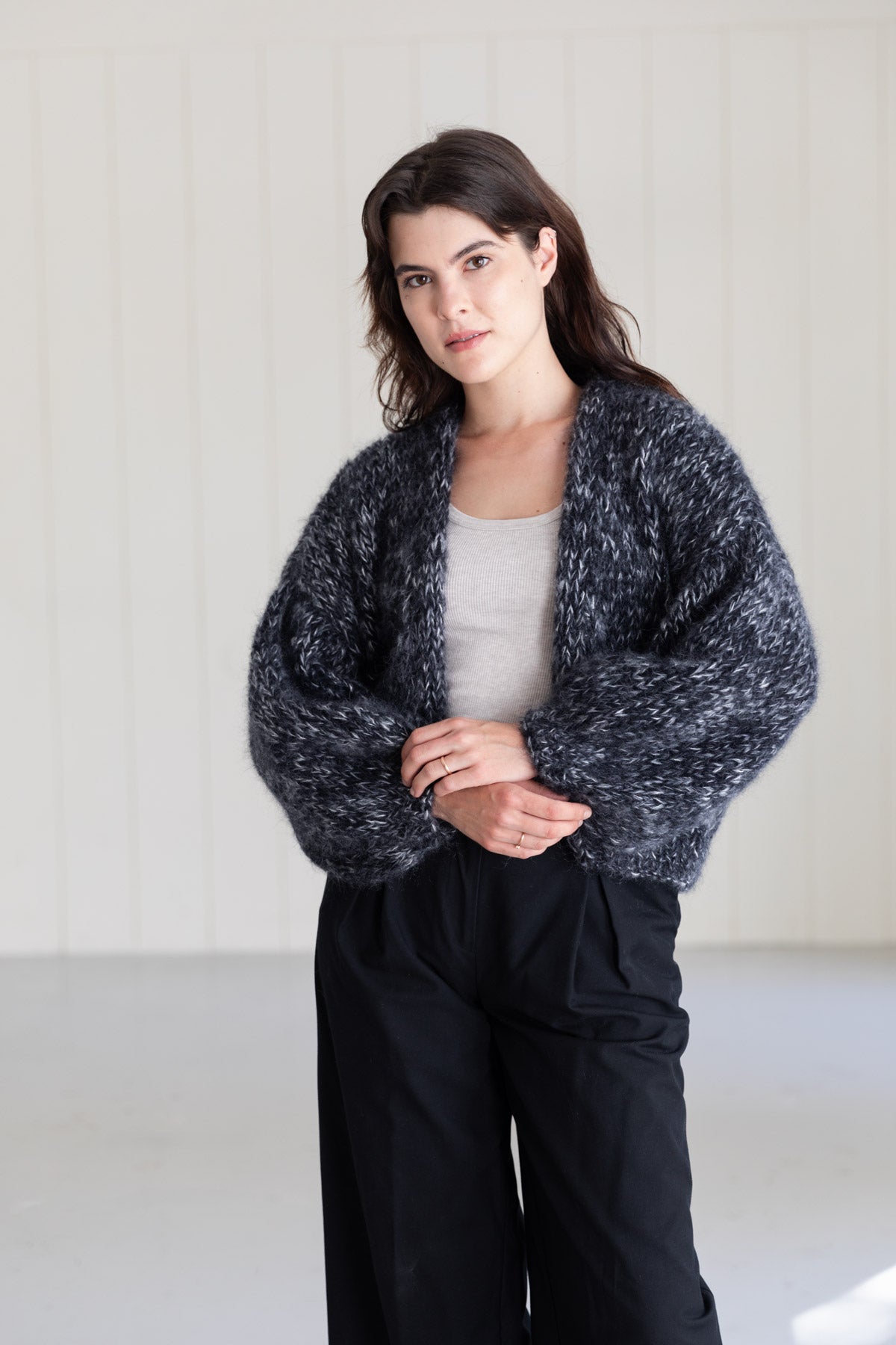 A woman wearing a chunky mohair cardigan with a marled black and white texture, worn over a light grey top and black trousers, set against a light-colored paneled wall.
