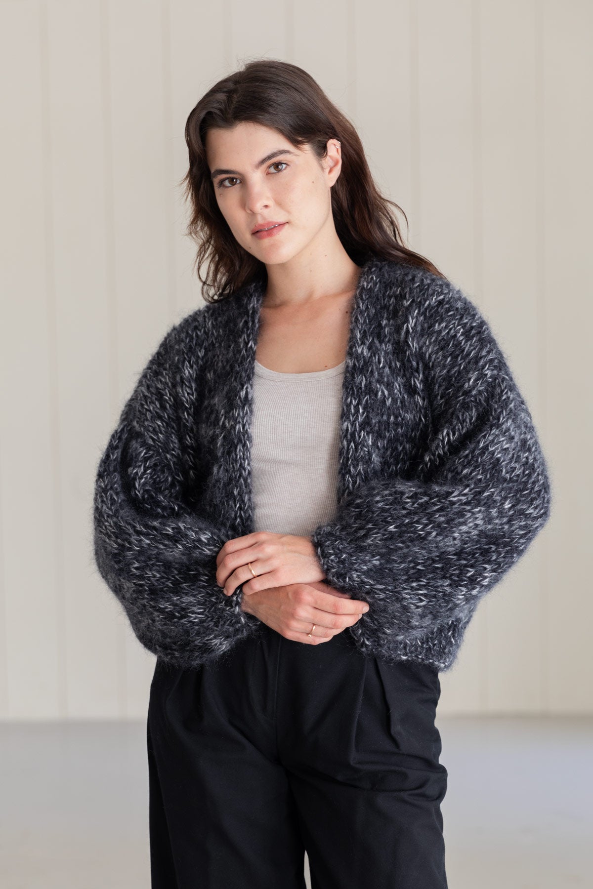 A woman wearing a chunky mohair cardigan with a marled black and white texture, worn over a light grey top and black trousers, set against a light-colored paneled wall.