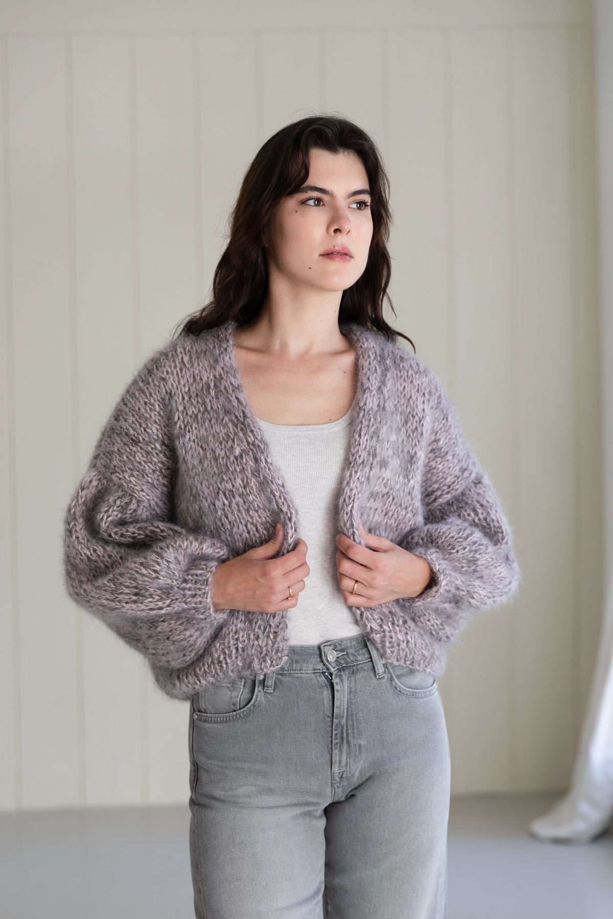 A woman wearing a chunky, lavender-grey mohair cardigan with balloon sleeves, styled over a white tank top and light-wash grey jeans.