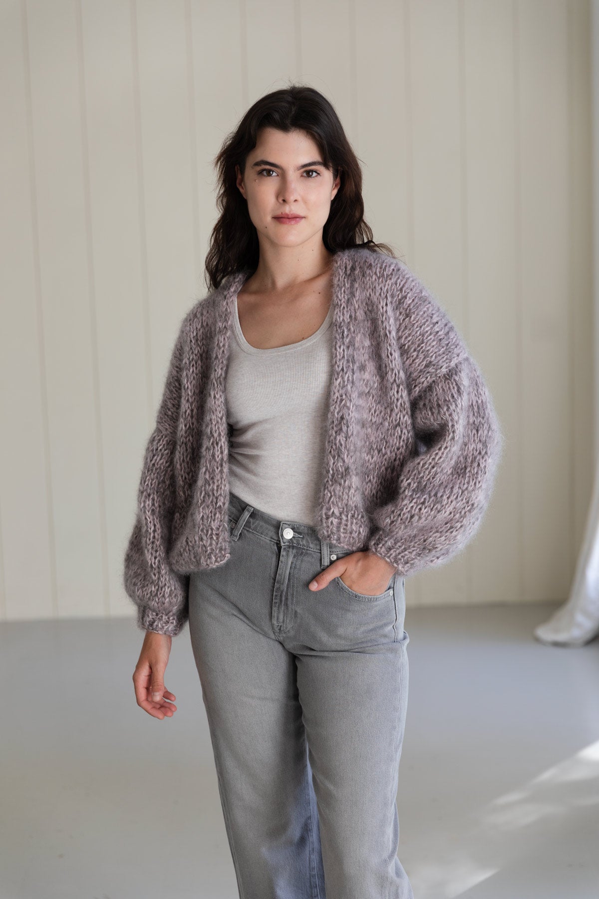 A woman wearing a bomber mohair cardigan in a dusty mauve-purple colour, worn over a light grey tank top and light-wash jeans.