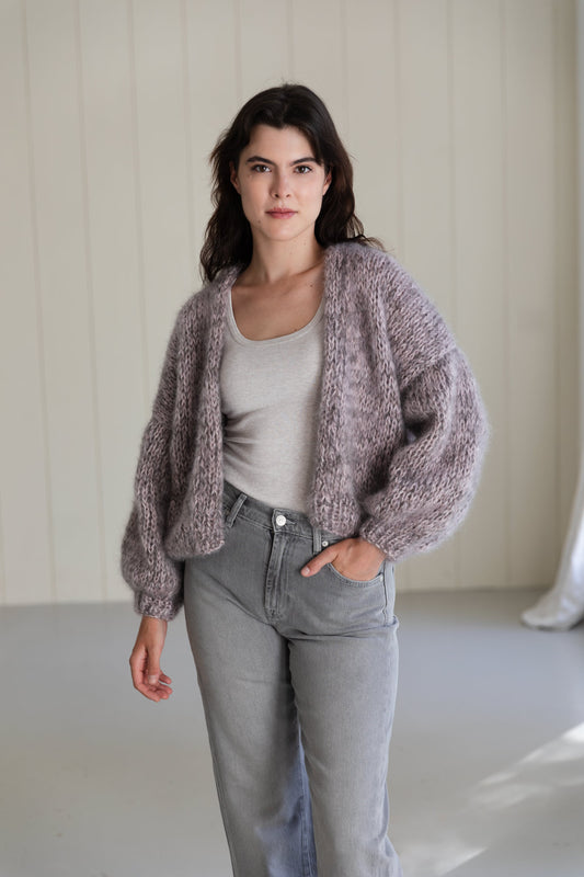 A woman wearing a bomber mohair cardigan in a dusty mauve-purple colour, worn over a light grey tank top and light-wash jeans.