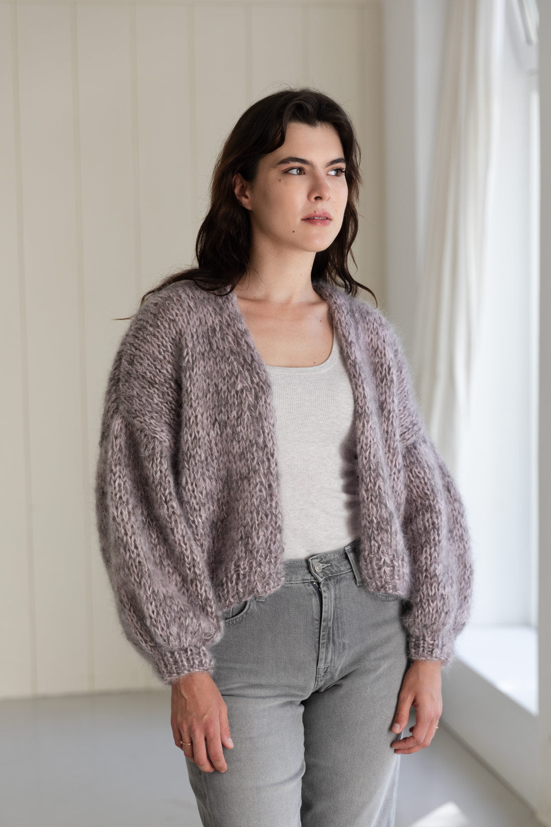 A woman wearing a chunky, fuzzy mohair cardigan in a dusty mauve-grey colour, styled over a light grey tank top and light-wash jeans.