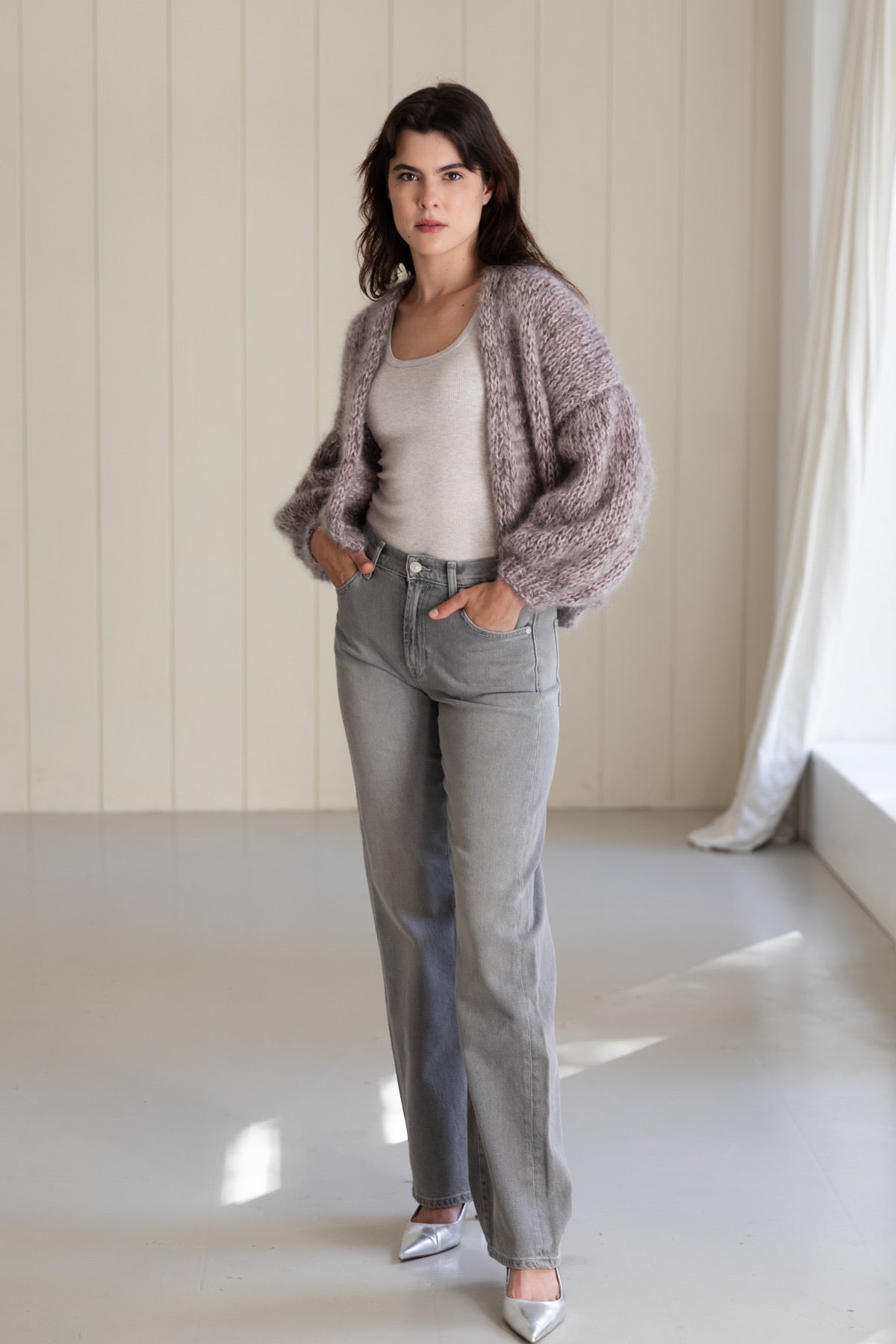 A woman wearing a bomber mohair cardigan over a light beige top, paired with grey jeans and metallic silver pointed shoes.