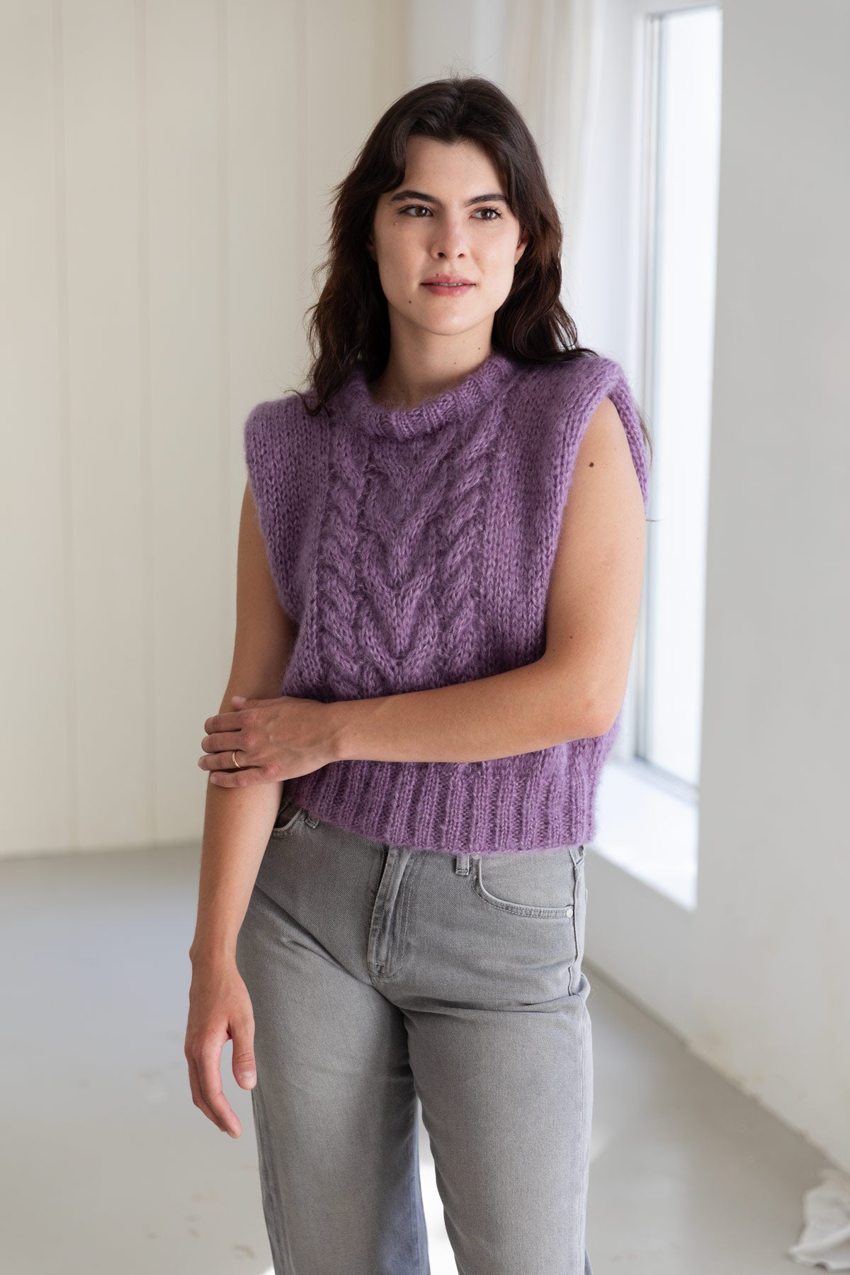 A woman wearing a lavender mohair sweater vest and light grey jeans, standing in a bright, minimalist room.