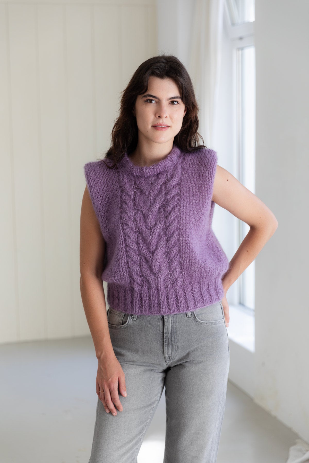 A woman wearing a sleeveless mohair sweater vest in purple, paired with light grey jeans, standing in a bright room.