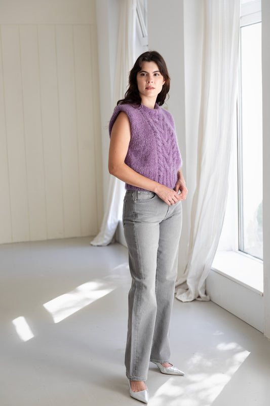 A woman stands in a bright, sunlit room wearing a lavender mohair sweater vest, light grey flared jeans, and silver pointed-toe flats.