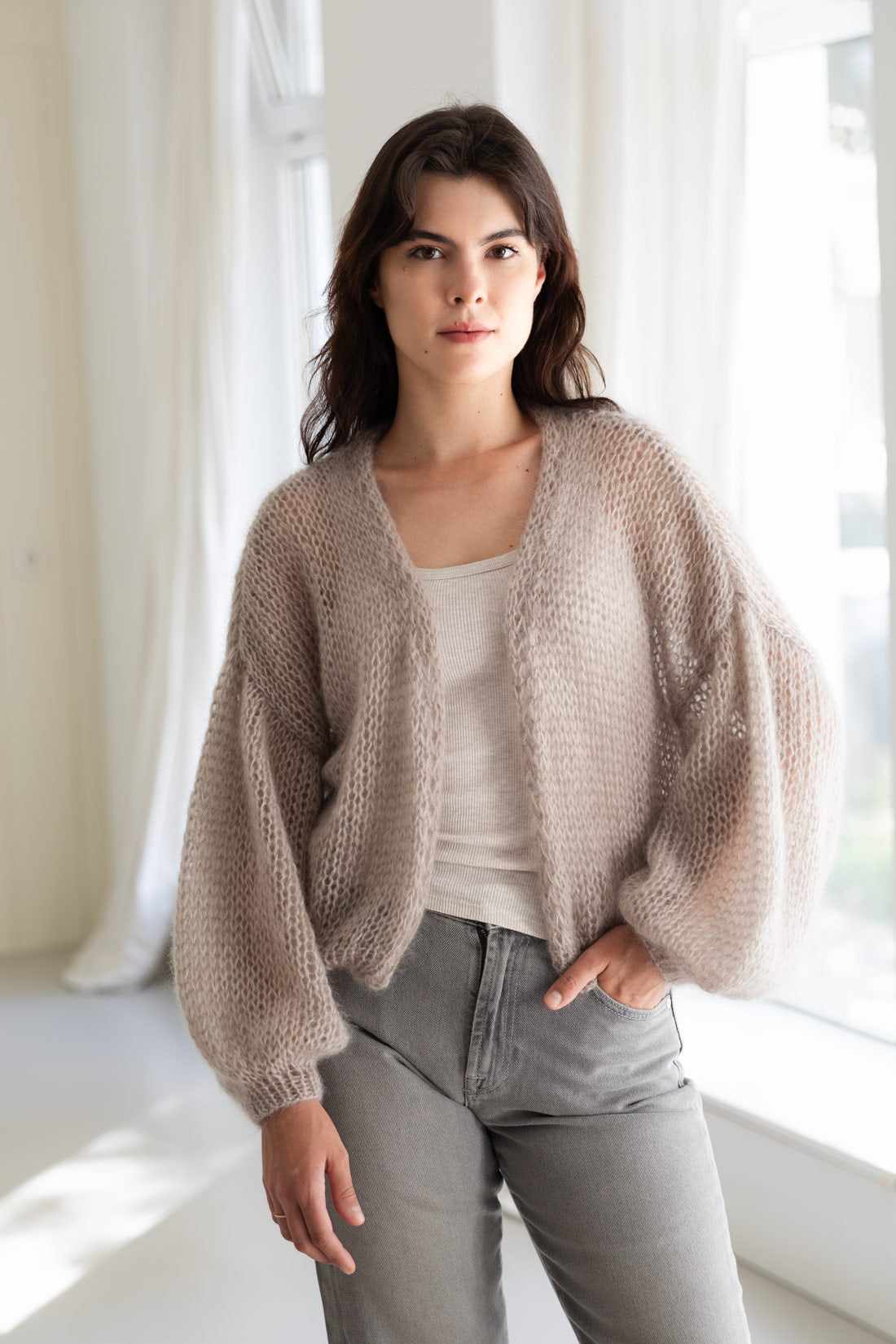 A woman wearing an airy mohair cardigan in a light taupe-beige colour, styled over a beige tank top and grey jeans.