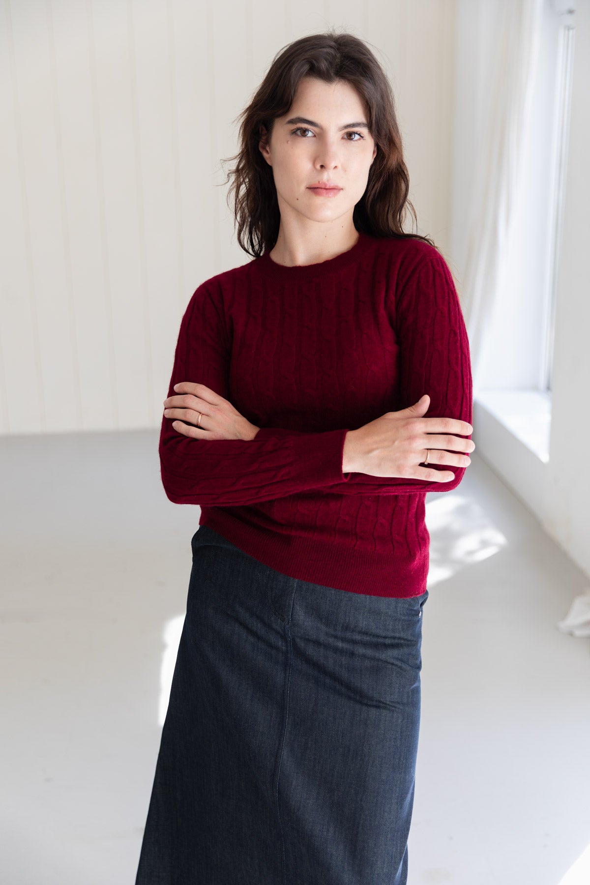 A woman wearing a dark red cable-knit cashmere sweater and a dark denim skirt, standing with arms crossed in a bright, minimalist room.