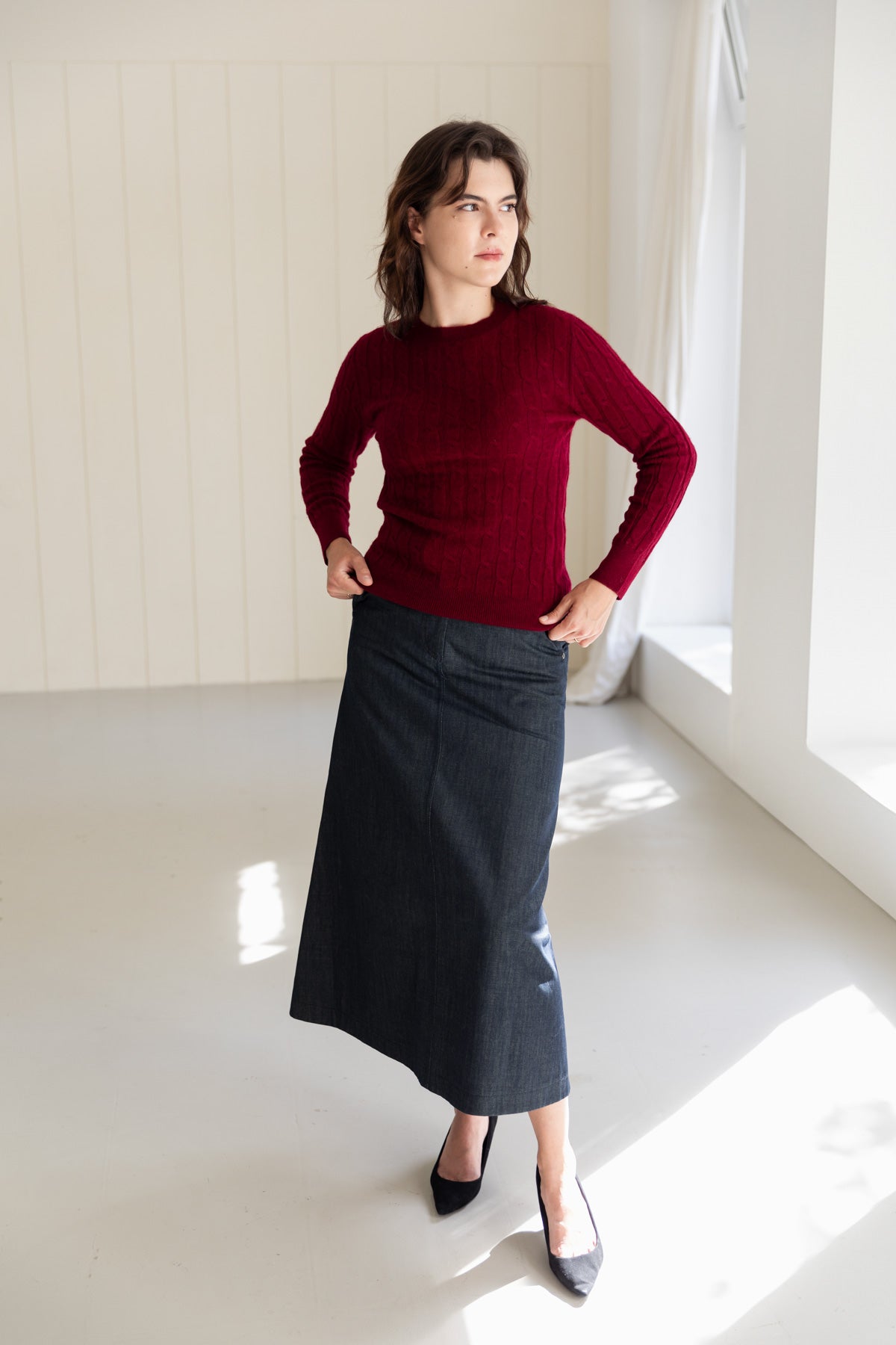 A woman stands in a bright, sunlit room wearing a dark red cable-knit cashmere sweater and a dark denim maxi skirt, paired with black pointed-toe flats.
